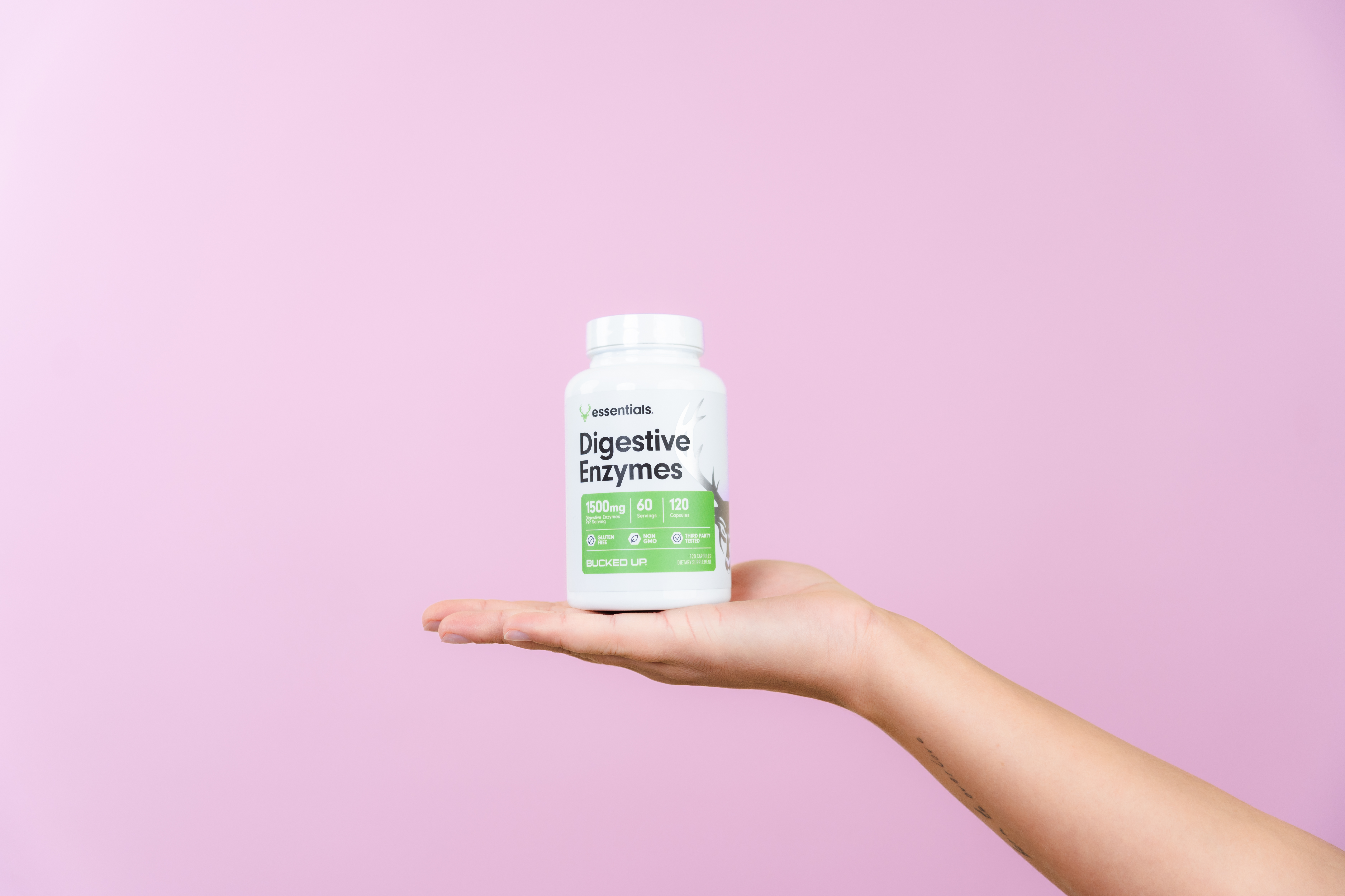 Hand holding Bucked Up Essentials Digestive Enzymes supplement bottle, labeled '1500mg,' '60 servings,' and '120 capsules,' against a light pink background.