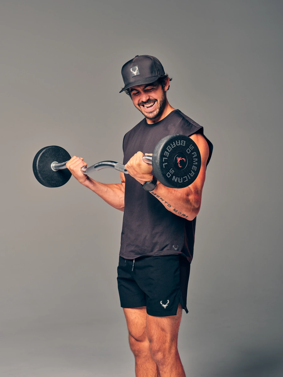 A young man with a beard and dark hair, wearing a black baseball cap with a logo, a sleeveless black athletic shirt, and black shorts with a white emblem, is lifting a black dumbbell with a silver handle in a studio setting with a plain gray background. He is smiling and looking down at the dumbbell, with his right arm bent at the elbow and his left arm slightly extended, showcasing his muscular build and a tattoo on his right forearm. The lighting highlights his face and upper body, creating a lively and energetic mood.