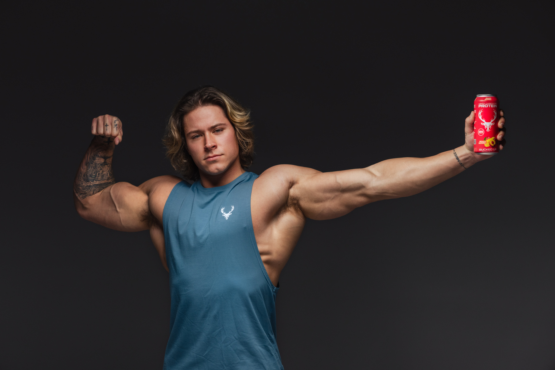 A muscular young man with shoulder-length wavy brown hair and light skin poses against a dark background, wearing a sleeveless blue athletic shirt with a white deer logo on the chest. He flexes his right bicep, which is tattooed with intricate black ink designs, and holds a red can of protein drink with a white deer logo and yellow accents in his extended left arm. His expression is confident and serious, with a slight squint and pursed lips, and the lighting highlights his defined muscles and facial features.