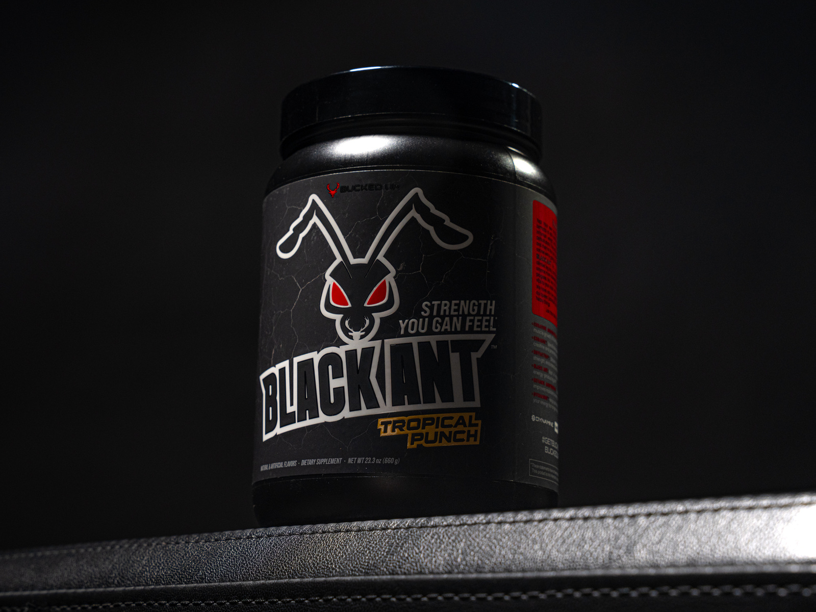 A sleek, black supplement container of Bucked Up Black Ant – Tropical Punch flavor is dramatically spotlighted against a dark background. The label features a bold, cracked-texture design with a stylized black ant logo that has glowing red eyes. The product slogan, “Strength You Can Feel,” is printed beside the ant’s head, conveying power and intensity. The overall composition highlights the product’s hardcore aesthetic and gym-performance appeal.