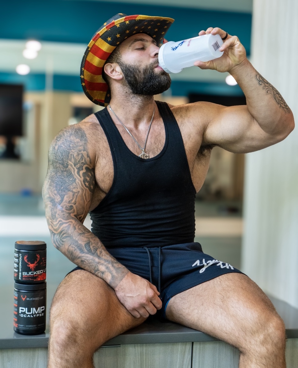 A muscular man with a full beard and tattoos on his left arm, wearing a black tank top, navy shorts, and a patriotic cowboy hat with stars and stripes, is sitting on a bench in a gym, drinking from a white shaker bottle. The background features gym equipment and a blue wall, with bright lighting highlighting his physique and the products on the bench, including two black containers labeled 'BUCKED UP PUMP' and 'BUCKED UP.' The overall mood is energetic and focused, with a professional fitness photography style.