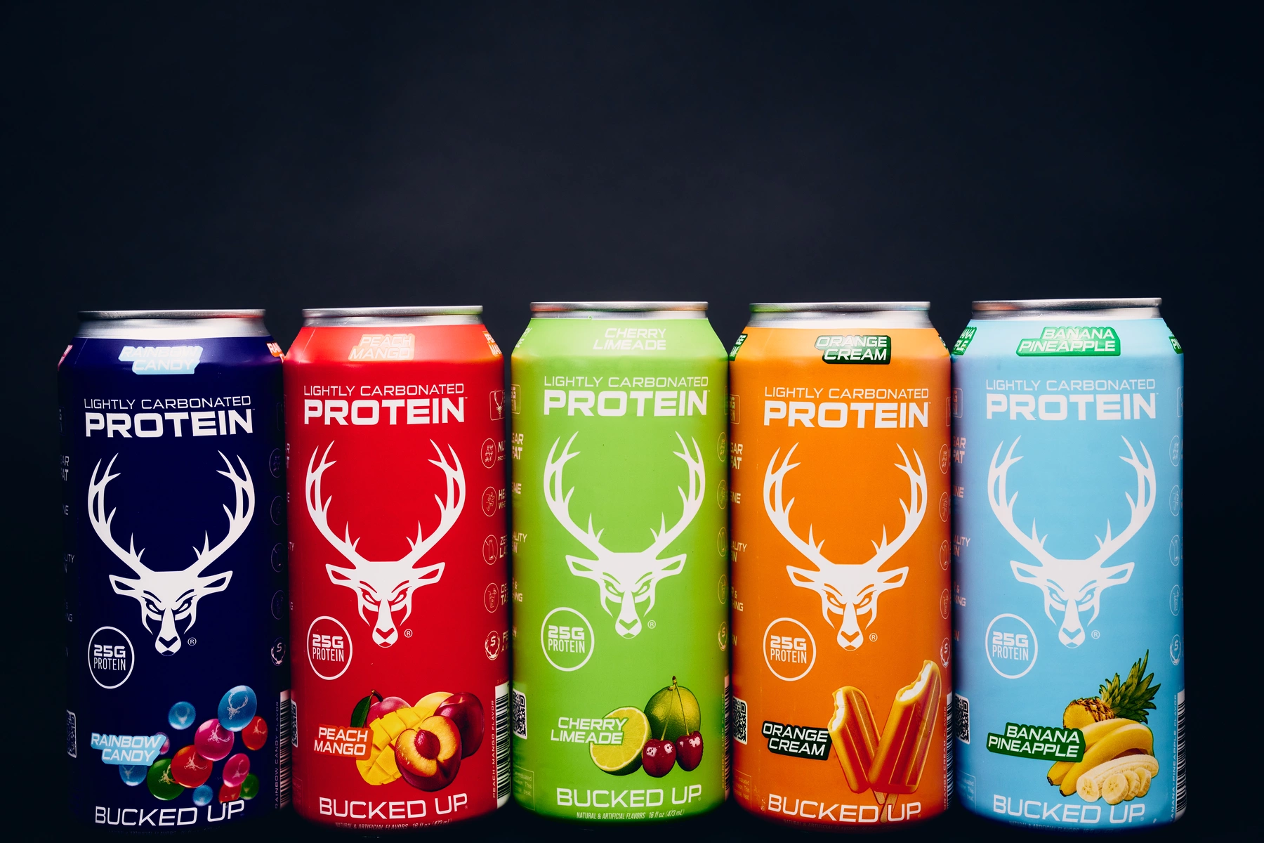Five cans of lightly carbonated protein drinks are arranged in a row against a dark background, each featuring a different vibrant color and flavor: rainbow candy in dark blue, peach mango in red, cherry limeade in green, orange cream in orange, and banana pineapple in light blue. The cans display a white deer head logo, flavor names at the top, and a small image of the fruit flavor at the bottom, with the text '25G PROTEIN' and 'BUCKED UP' on each. The lighting highlights the metallic sheen of the cans, emphasizing their bright colors and sleek design.