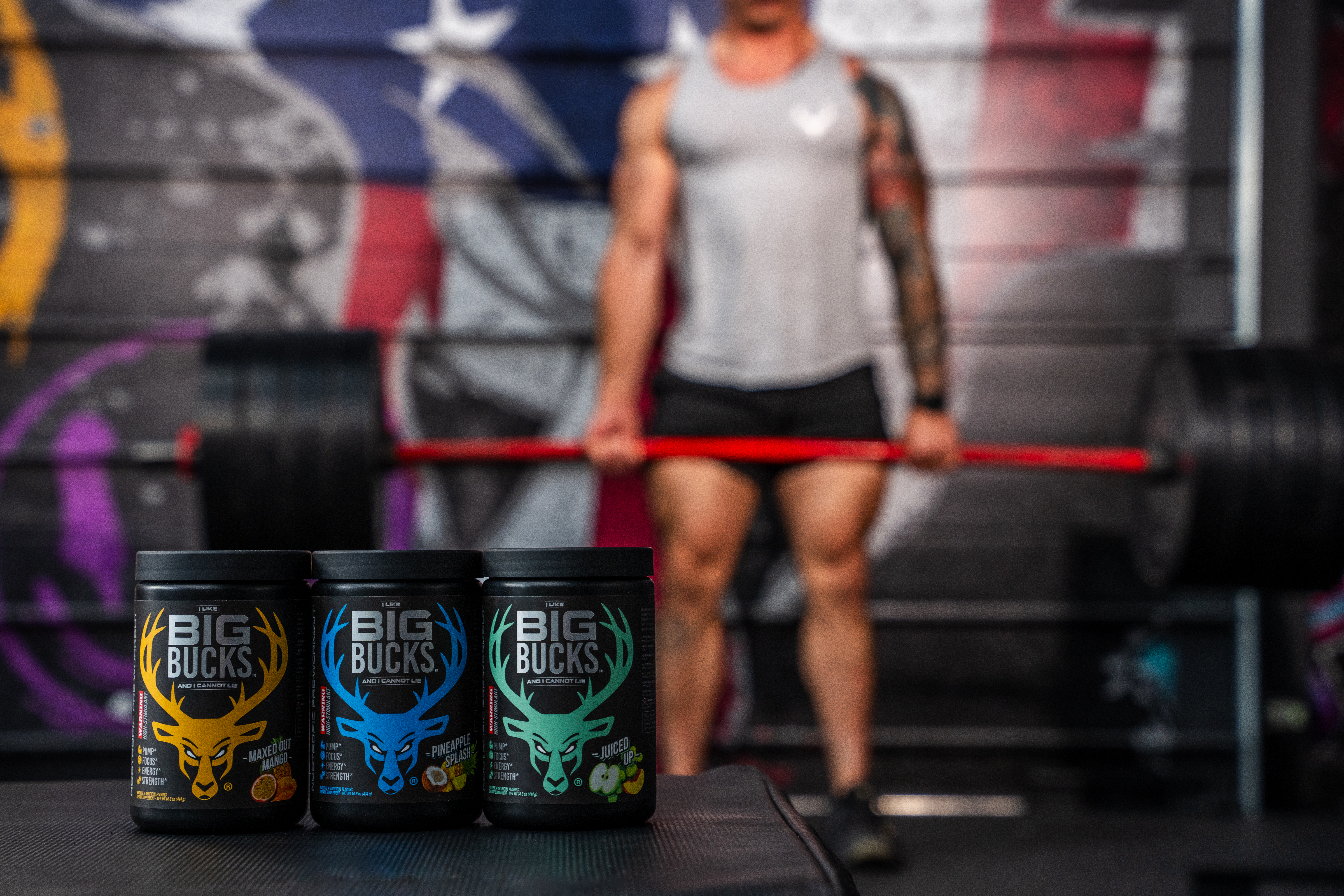 Three tubs of BIG BUCKS pre-workout supplements—Maxed Out Mango, Pineapple Splash, and Juiced Up—displayed in front of a muscular man deadlifting a loaded barbell in a gym with a patriotic mural in the background.