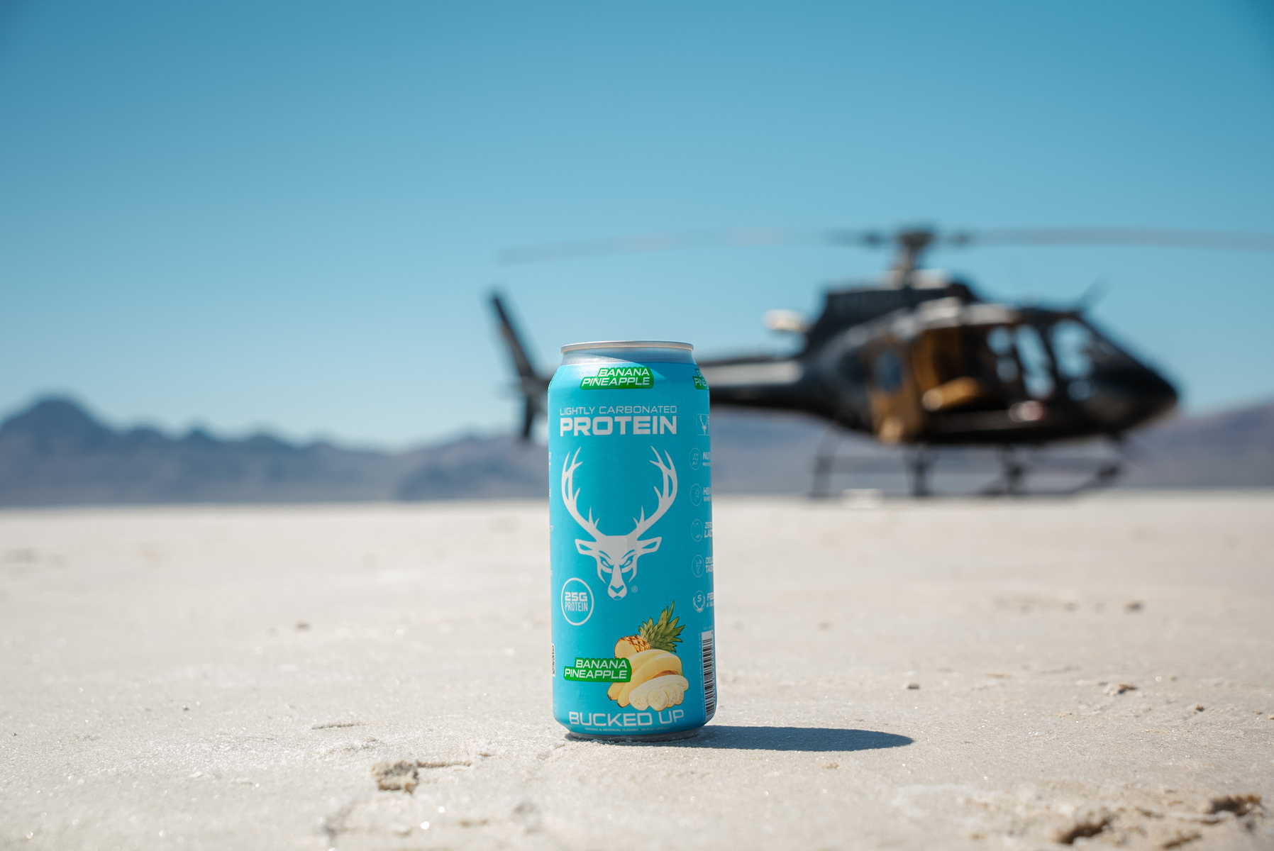 A bright blue can of lightly carbonated banana pineapple protein drink with a white deer head logo and green accents, standing upright on a flat, sandy desert surface with a clear blue sky in the background. In the blurred background, a black helicopter with gold accents is parked on the sand, with mountains visible in the distance. The scene is well-lit with natural sunlight, casting soft shadows, and the overall mood is adventurous and energetic, suitable for outdoor or sports branding.