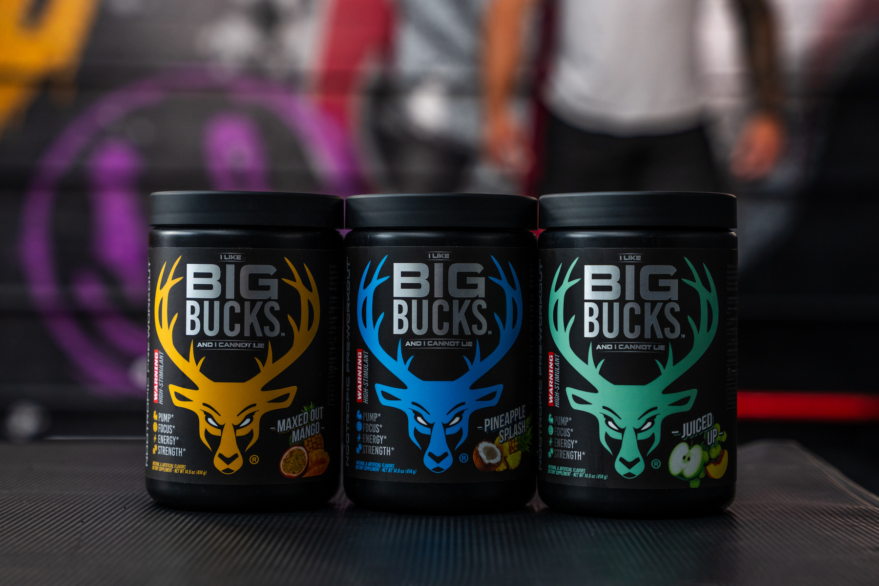 Three black containers of dietary supplements labeled 'Big Bucks' are arranged side by side on a textured black surface, with the yellow, blue, and green logos featuring stylized deer antlers. The labels indicate flavors: Maxed Out Mango, Pineapple Splash, and Juiced Up, with small icons of fruits and key benefits such as pump, focus, energy, and strength. The background is blurred, showing a graffiti wall with purple and pink accents and a person wearing a gray shirt and black pants standing behind the containers, with soft, directional lighting highlighting the containers and creating subtle shadows.