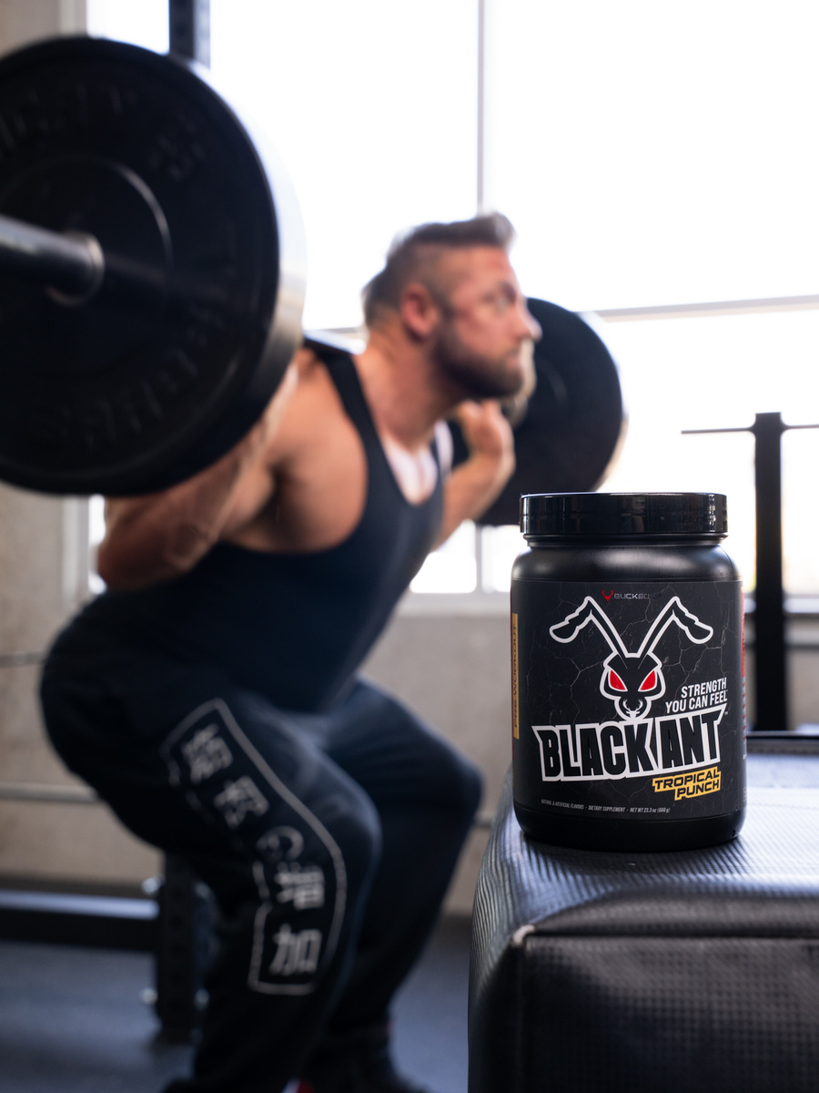 In a bright weight room, a powerful athlete sinks into a barbell back squat, muscles taut and posture dialed in. The camera throws the lifter softly out of focus, placing crisp attention on a matte-black supplement jar perched on a bench. The label features a fierce ant icon with red eyes above the bold name BLACK ANT, signaling grit and intensity. Framed by steel uprights and sunlight, the composition uses shallow depth to connect product and performance without distraction. It’s a lifestyle shot that sells the feeling of focus, discipline, and strength that serious training demands, before the next rep.