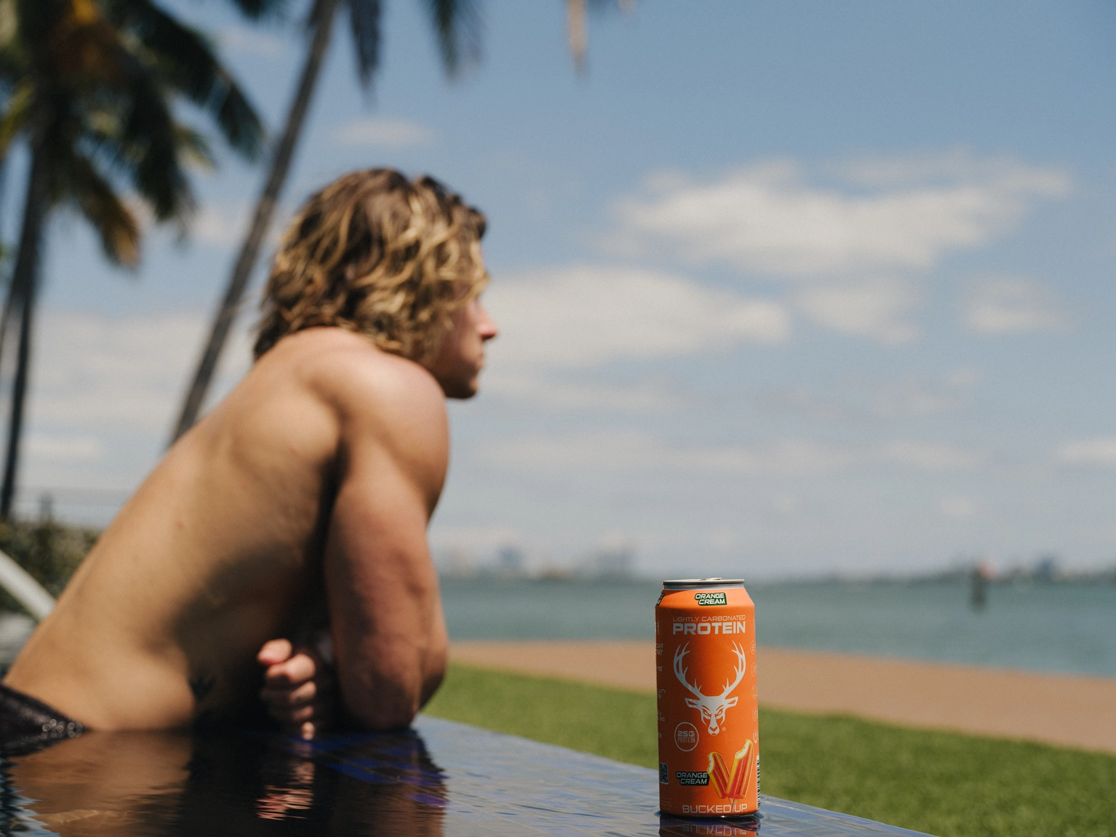 A young man with wavy, shoulder-length blond hair and light skin is leaning on the edge of a pool, gazing into the distance with a contemplative expression. He is shirtless, revealing a toned physique, and is wearing dark swim trunks. The background features a clear blue sky with a few scattered clouds, tall palm trees, and a distant view of the ocean with some buildings on the horizon. In the foreground, an orange can of protein drink with a deer logo and the words "Lightly Carbonated Protein" and "Orange Cream" is placed on the poolside ledge, reflecting in the water. The scene is bright, sunny, and has a relaxed, summery vibe.