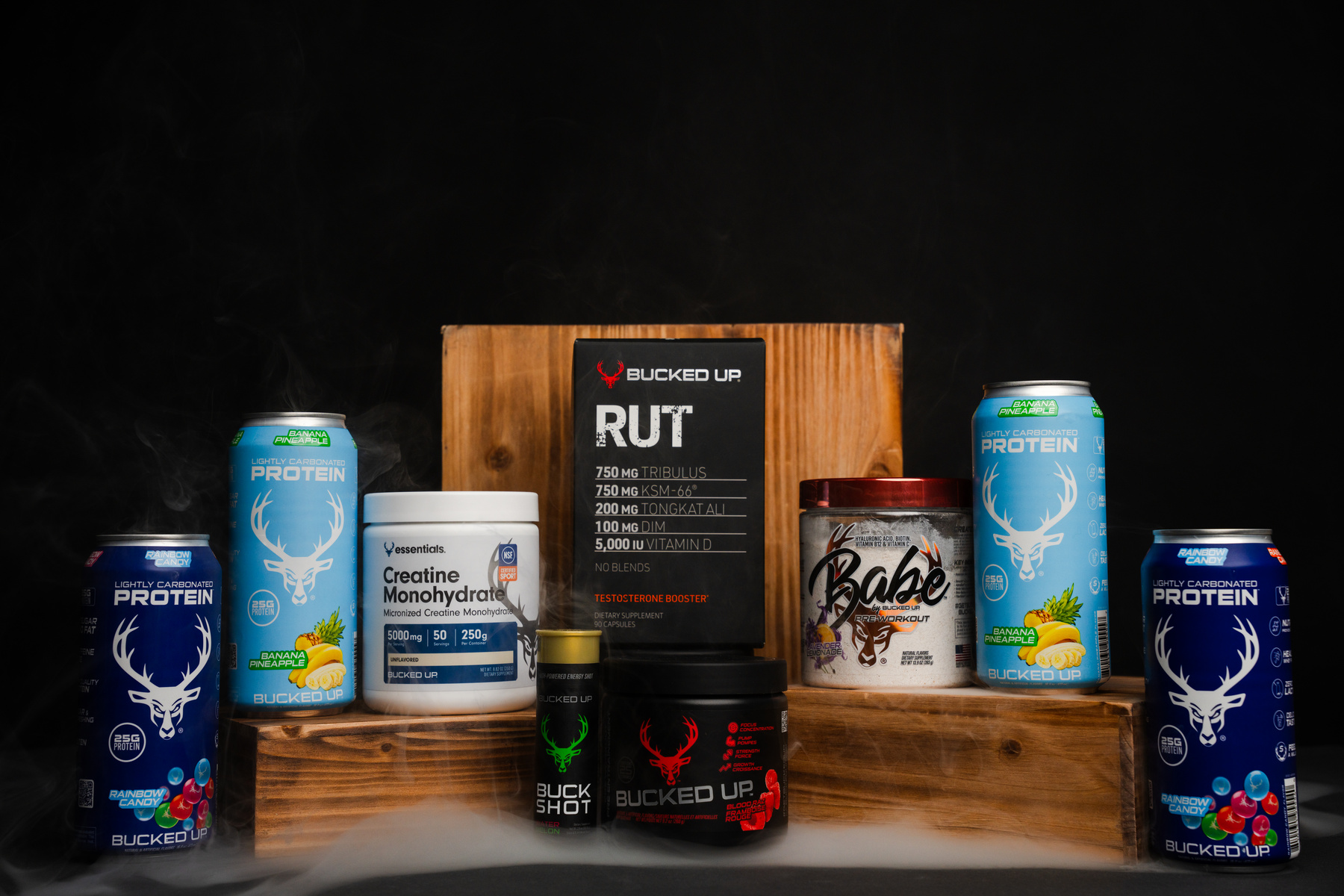 A display of fitness supplements arranged on a wooden platform against a black background, including two blue cans of rainbow candy flavored protein with a white deer logo, a white container of creatine monohydrate, a black container of Bucked Up BCAA with a green deer logo, a red-lidded container of Babe pre-workout, a black box of Bucked Up RUT testosterone booster, and a yellow capsule of Bucked Up energy shot, with steam rising from the cans, highlighting a professional, energetic, and health-focused mood with bright, vivid colors and clear branding.