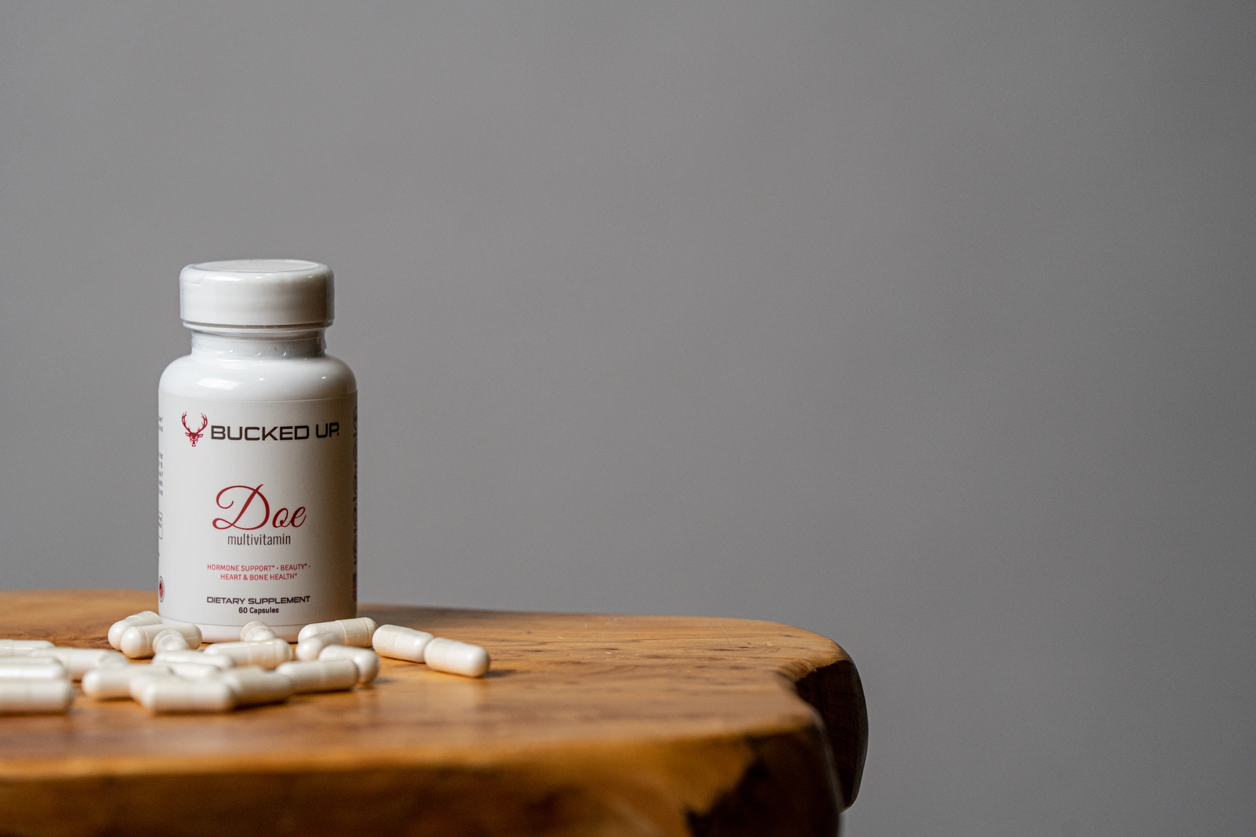 A white plastic bottle labeled 'Bucked Up Doe multivitamin' with a red deer logo, placed on a rustic wooden surface with several white capsules scattered around it, set against a plain gray background. The bottle is upright, with a white screw cap, and the label indicates it is a dietary supplement for hormone support, beauty, heart, and bone health.