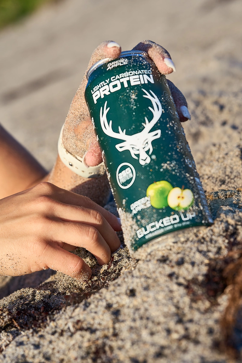 A person with sandy fingers holds a can of lightly carbonated protein drink labeled 'BUCKED UP' with a deer head logo and green apple flavor, partially buried in sand on a beach, with a blurred sandy background. The can is dark green with white text and graphics, and the person's nails are painted white with a hint of sand on their fingers. The scene is brightly lit with natural sunlight, creating shadows and highlights on the can and sand, evoking a casual, outdoor, beach vibe.