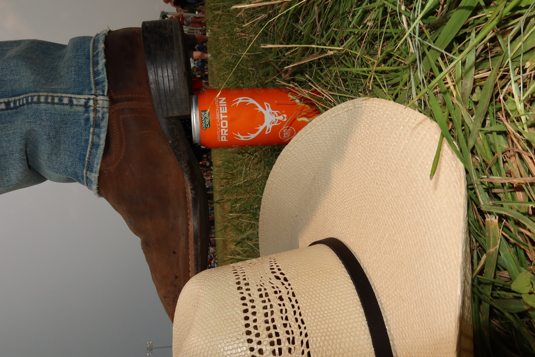 A person wearing blue jeans and brown leather boots is standing on a grassy field, with the boot pressing down on an orange protein drink can featuring a white deer head logo. In the foreground, a beige straw hat with a black band rests on the grass, and in the background, there are blurred figures of people and a cloudy sky, suggesting an outdoor event or gathering. The image is taken from a low angle, emphasizing the boot and can, with natural lighting and a casual, outdoor atmosphere.