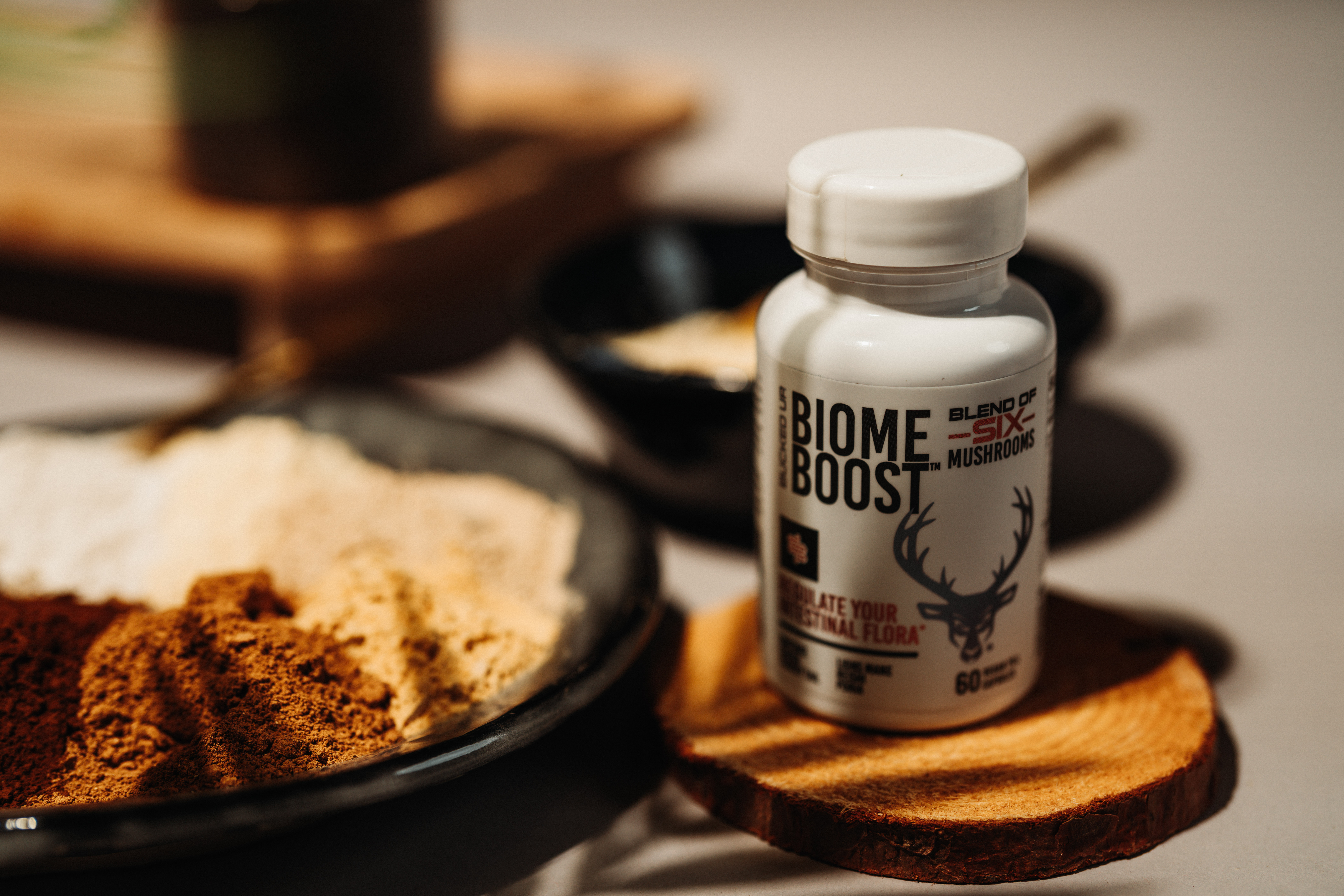 Close-up image of Bucked Up Biome Boost supplement bottle on a wooden coaster, labeled 'Blend of Six Mushrooms' and 'Stimulate Your Intestinal Flora,' beside assorted mushroom powders.