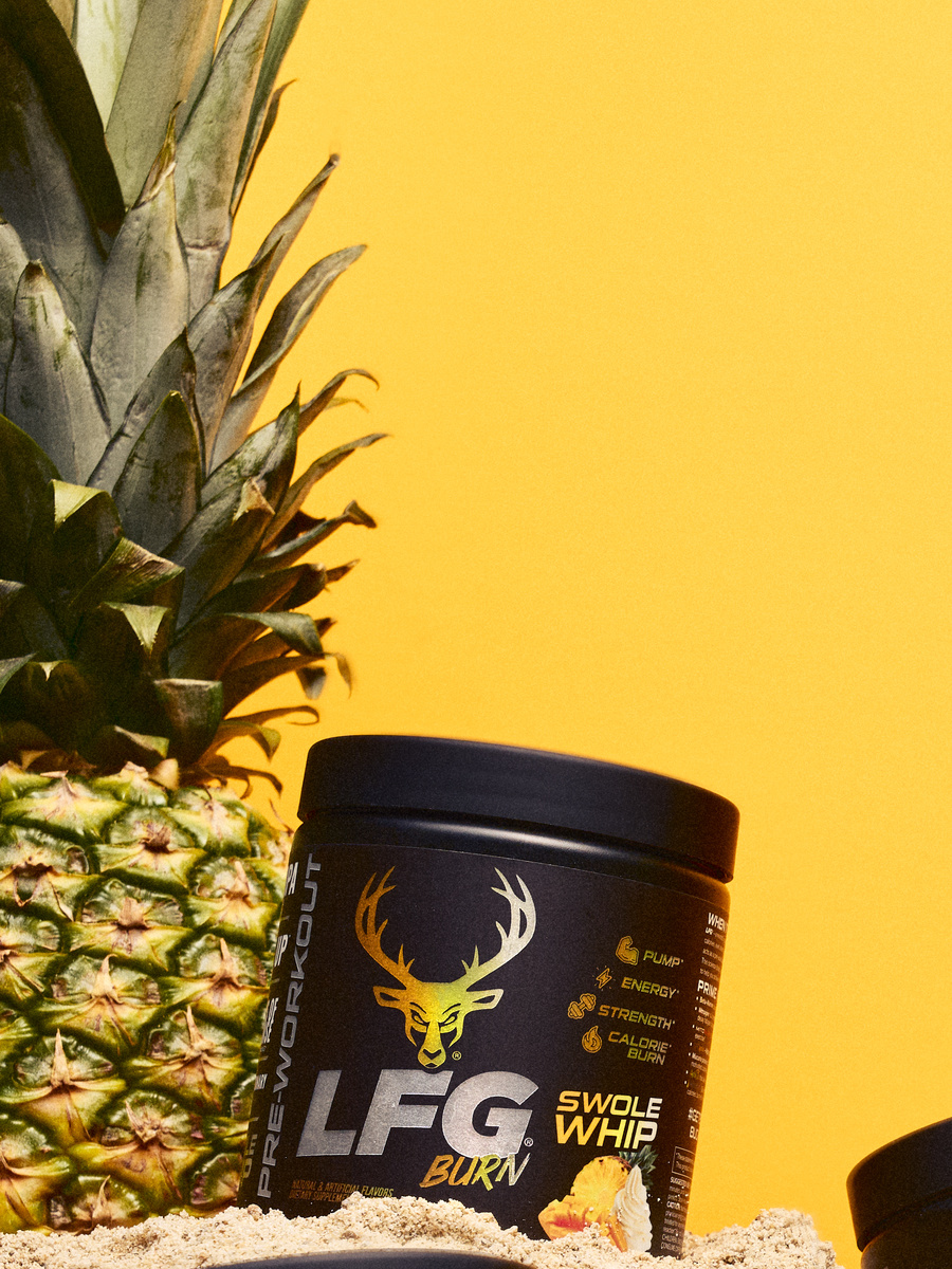 A black container of LFG Burn supplement with a yellow deer logo and white text, placed on sandy ground next to a ripe pineapple with spiky green leaves, set against a bright yellow background. The container features icons indicating benefits such as pump, energy, strength, and calorie burn, with a scoop of yellow powder and whipped cream illustration on the label. The composition is close-up, with the pineapple partially visible on the left, and the container centered, illuminated by soft, even lighting that highlights the product details and vibrant colors.