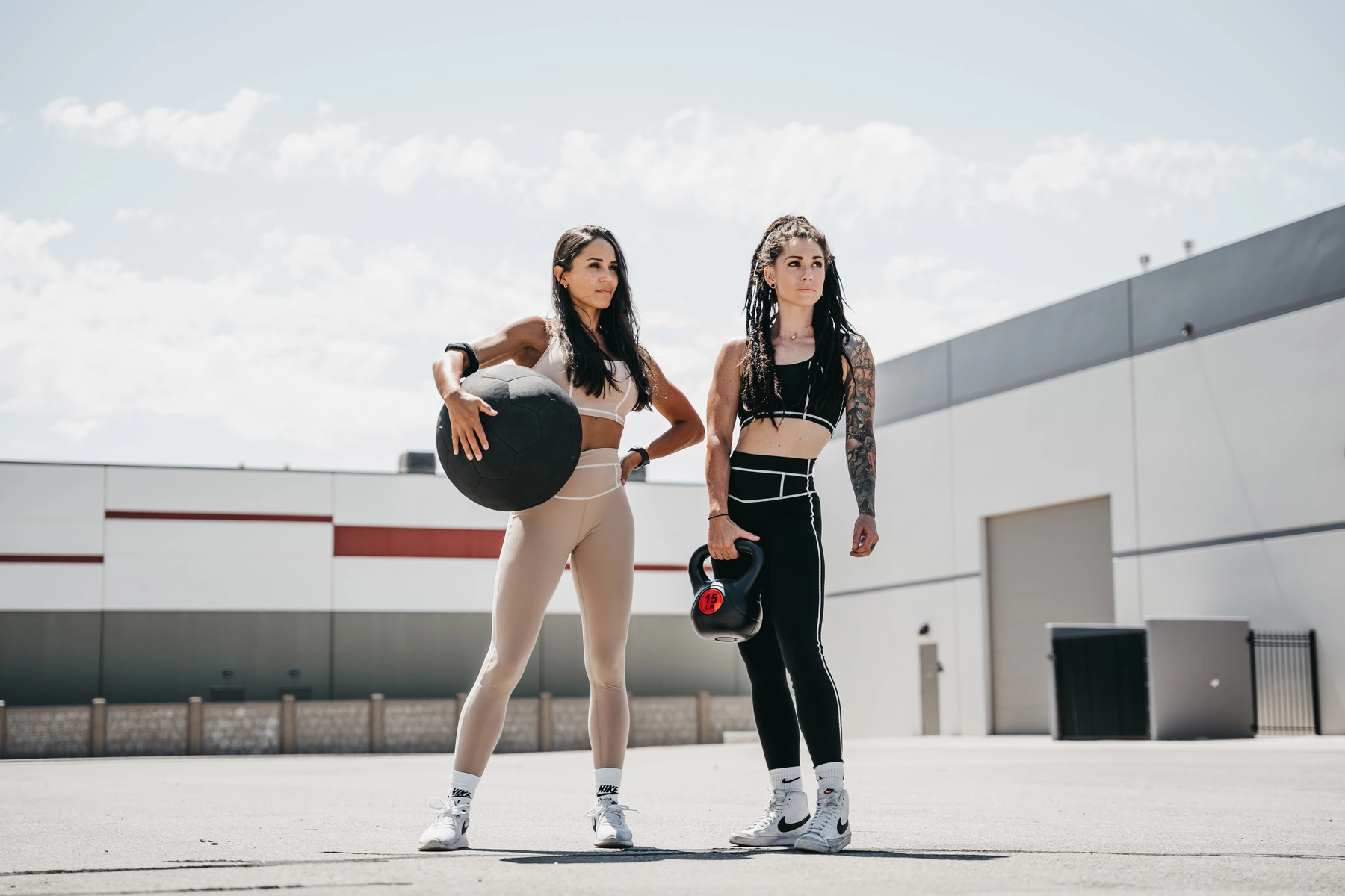 Two fit young women with athletic builds stand outdoors on a sunny day in front of a modern industrial building, dressed in matching black and beige workout outfits with white accents, holding fitness equipment; the woman on the left holds a large black medicine ball, while the woman on the right holds a black kettlebell with a red label, both wearing white Nike sneakers and socks, with the woman on the right having long dreadlocks and visible tattoos on her left arm, and the woman on the left having long dark hair, with a clear blue sky and scattered clouds in the background, captured from a low angle emphasizing their confident stances, with bright natural lighting highlighting their determined expressions.