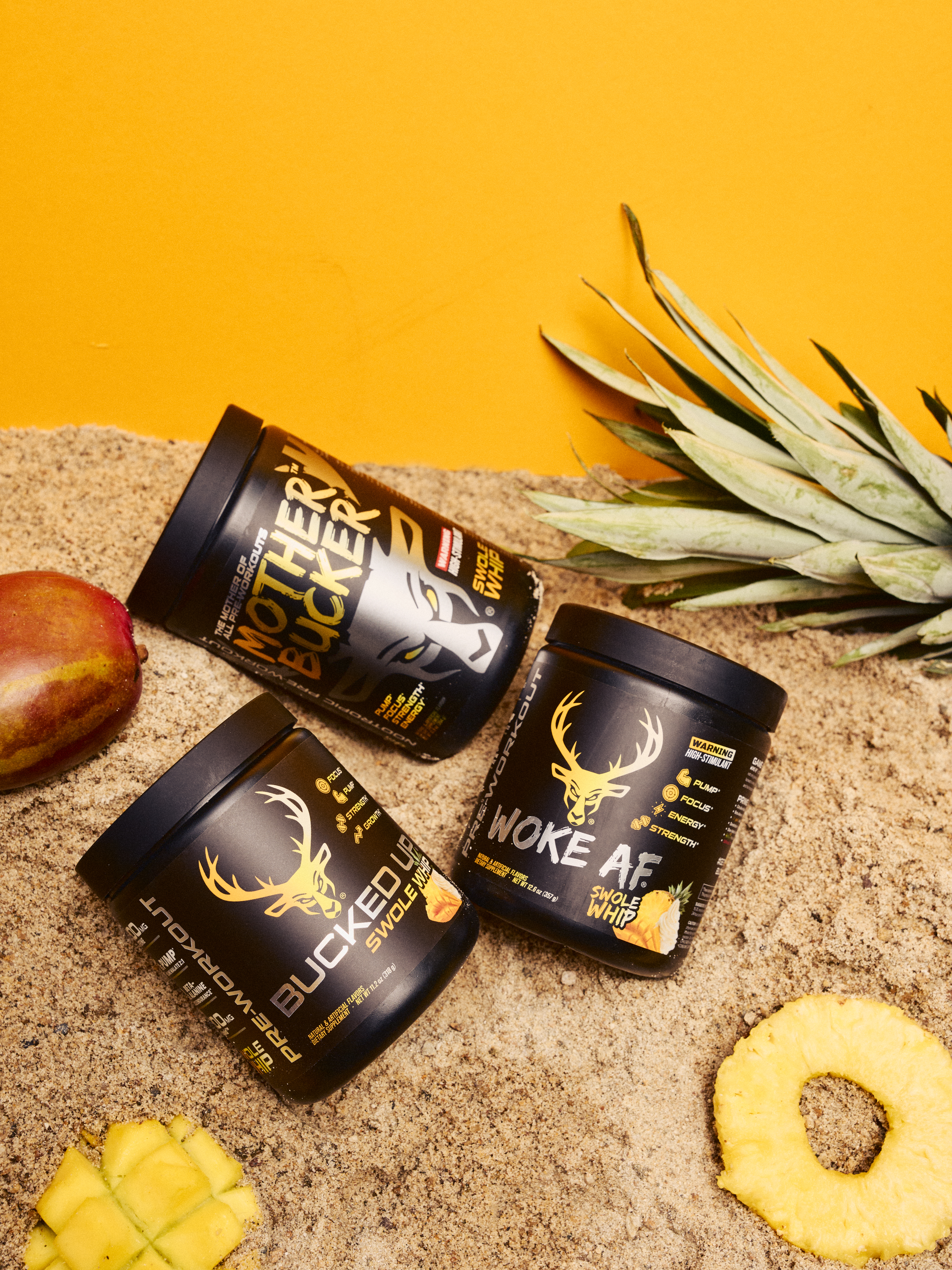Three black containers of dietary supplements with gold and white branding, featuring a deer head logo, are placed on a sandy surface with a yellow background. One container is labeled "Mother Buckery" with a stylized wolf face, while the other two are labeled "Bucked Up" and "Woke AF" with pineapple illustrations. The scene includes a pineapple crown, a pineapple ring, a mango, and a small piece of pineapple, arranged around the containers. The lighting is bright and even, emphasizing the vibrant yellow background and the contrast of the black containers, creating a lively, energetic mood suitable for a fitness or supplement advertisement.