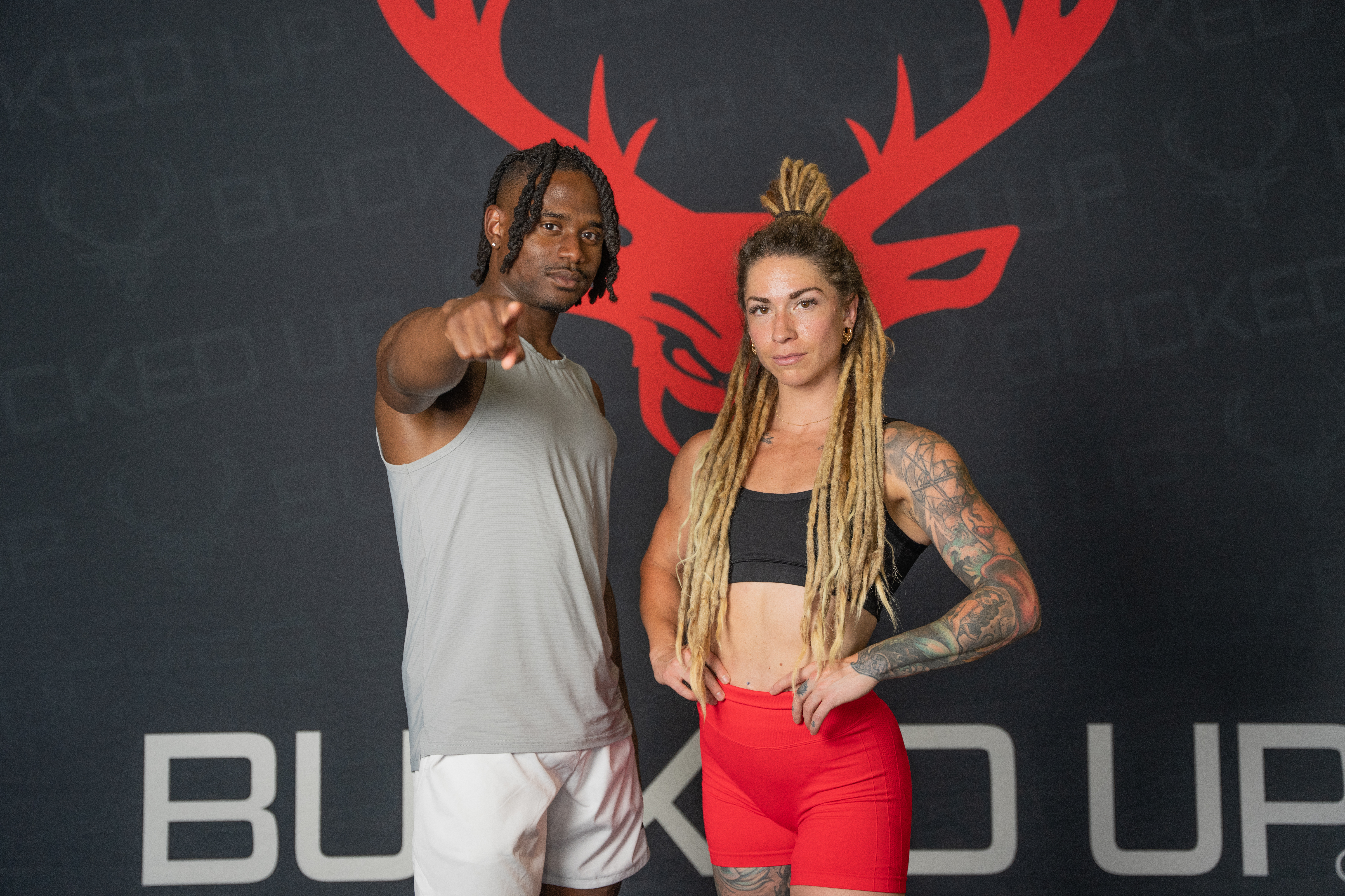 Two athletic individuals confidently posing against a Bucked Up branded backdrop featuring a prominent red deer logo, one pointing directly at the camera.