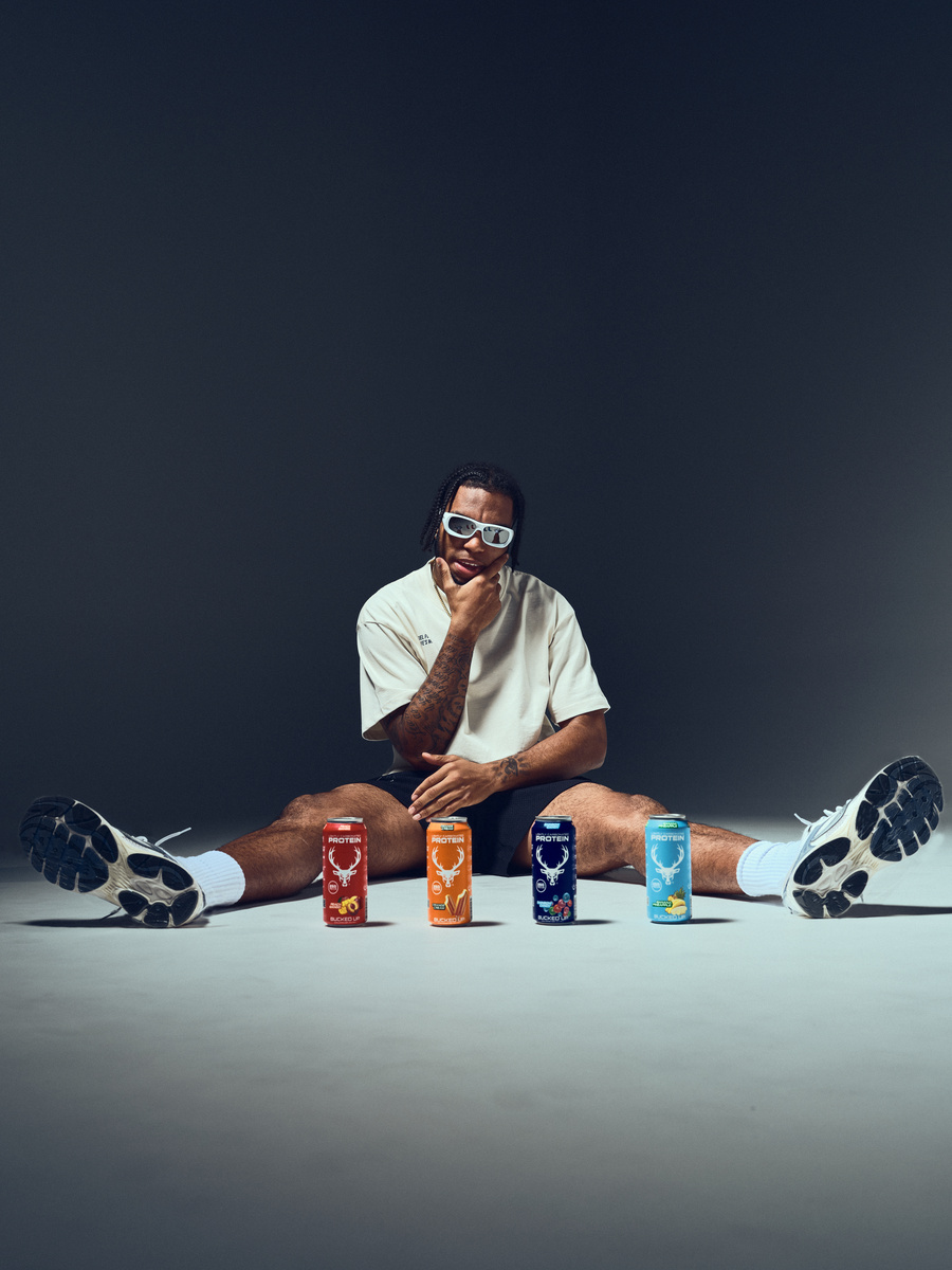 A young man with dark skin, dreadlocks, and tattoos on his arms is sitting on the floor with legs spread wide apart, wearing a loose white t-shirt, black shorts, white socks, and black and white athletic shoes. He is wearing large white sunglasses and has a thoughtful expression, with one hand resting on his chin. In front of him are four colorful cans of protein drinks arranged in a row, each with a different color and design, set against a plain, dark background with soft lighting highlighting his face and the cans.