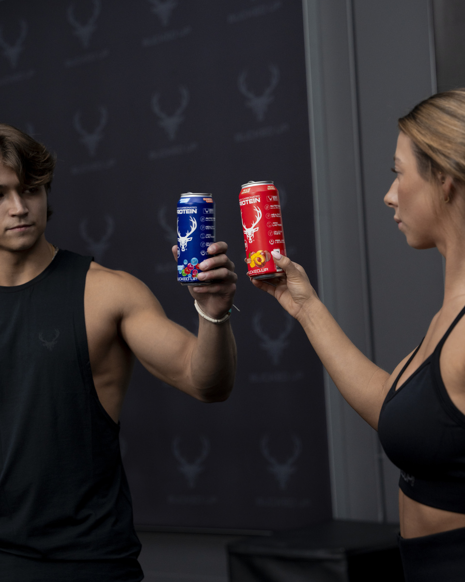 A muscular young man with wavy brown hair, wearing a black sleeveless athletic shirt with a subtle logo, is holding a blue can of protein drink in his right hand, extending it towards a woman with light brown hair tied back, wearing a black sports bra, who is holding a red can of protein drink in her right hand, both standing in front of a dark backdrop with a repeated logo pattern, under soft, focused lighting that highlights their faces and the cans, creating a professional, energetic atmosphere suitable for a fitness or supplement promotion.