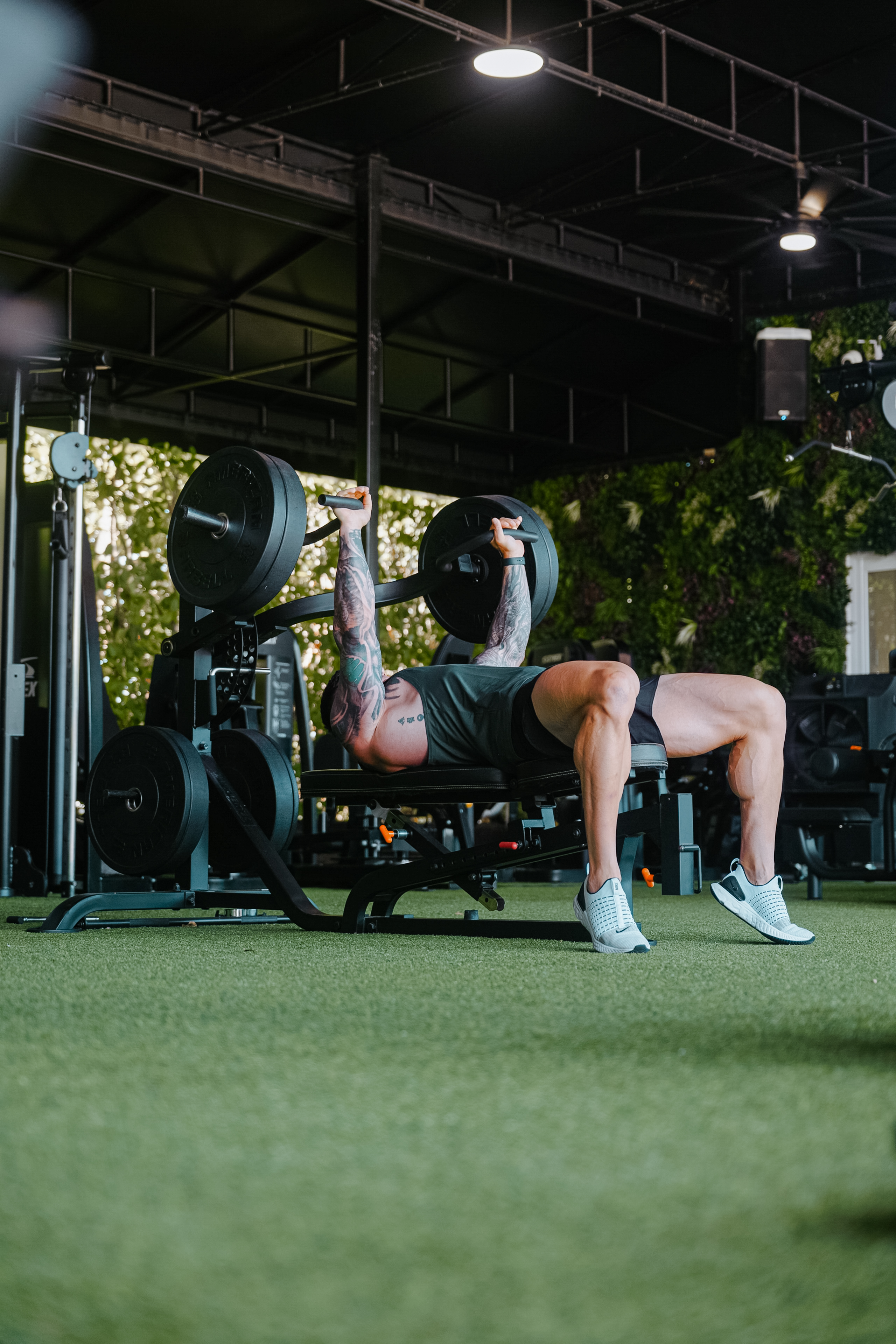“Athlete performing bench press with heavy weights wearing Bucked Up sleeveless gym shirt and athletic shorts – strength training and fitness apparel photo.”