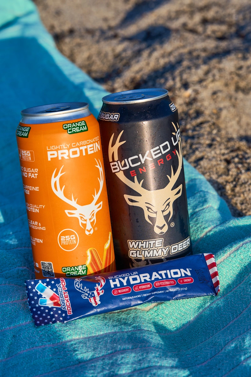Two energy drink cans, one orange with a white deer head logo and the words "Lightly Carbonated Protein" and "Orange Cream," and the other black with a white deer head logo and the words "Buckled Up Energy" and "White Gummy Deer," are placed on a blue towel on sandy ground, with sunlight casting shadows and highlighting the metallic surfaces of the cans.