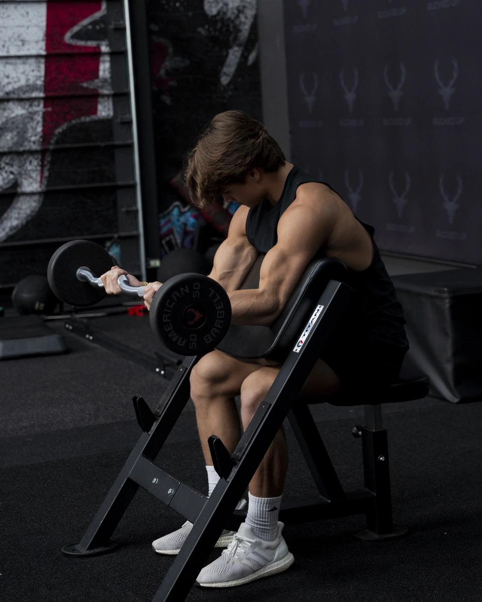 A young male athlete with a muscular build, light skin, and brown hair, is seated on a black workout bench in a gym, performing a bicep curl with a silver dumbbell labeled 'Dumbbell 50' in black. He is wearing a sleeveless black athletic shirt, black shorts, white Nike socks, and white athletic shoes. The background features a dark wall with a repeating logo pattern and a graffiti-style mural with red, black, and white colors. The gym environment is dimly lit with focused lighting highlighting the athlete's muscles and the equipment. The composition is a side view, emphasizing his arm muscles and concentration during the exercise.