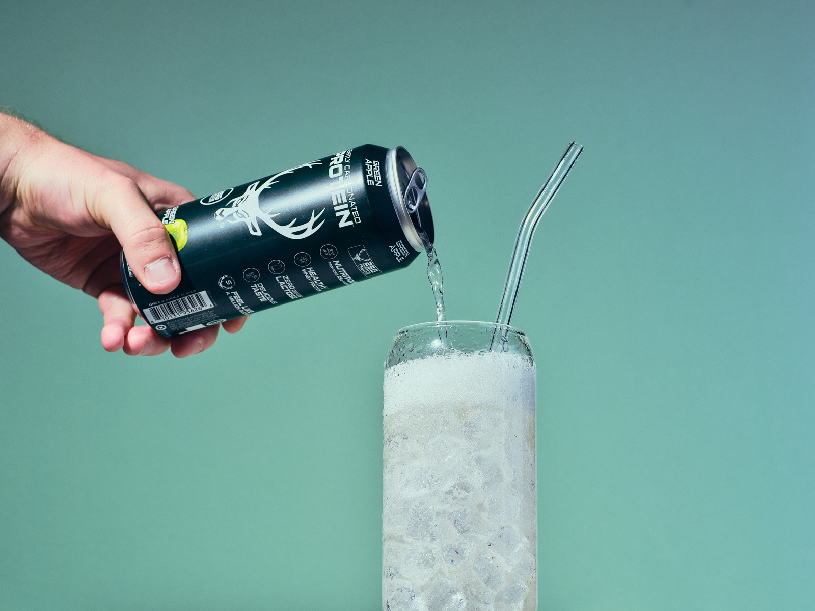 A hand is pouring a black can of Green Apple flavored sparkling water into a tall glass filled with ice, with a clear glass straw inside. The can features a white deer head logo and various icons and text, including the flavor and brand details. The background is a smooth, gradient teal color, and the lighting highlights the condensation on the can and the ice in the glass, creating a fresh and vibrant mood. The composition is focused on the pouring action from a side angle, emphasizing the contrast between the dark can and the light, icy drink.