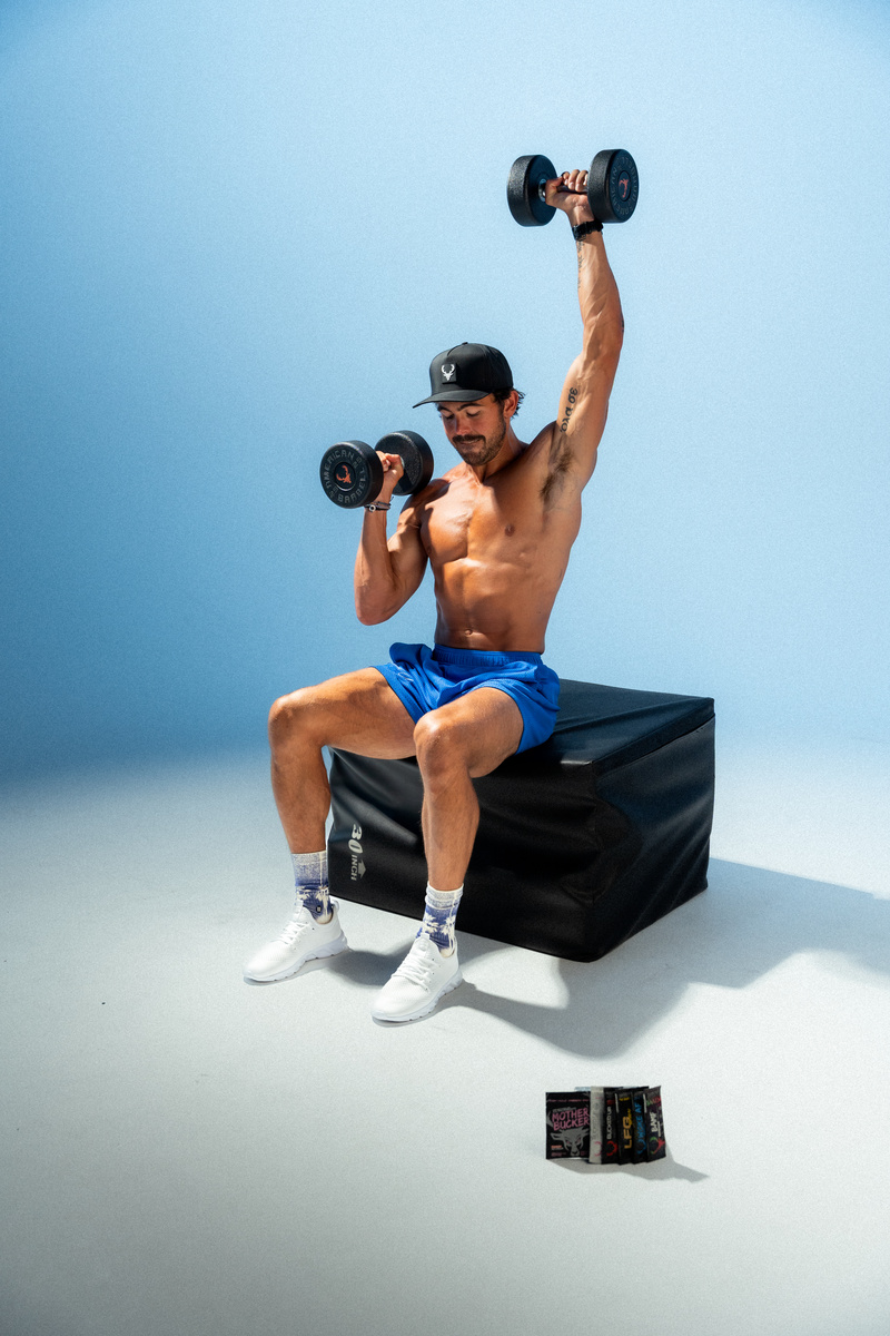 A muscular man with a beard, wearing a black cap with a white logo, blue shorts, white sneakers, and patterned socks, is seated on a black padded box against a plain light blue background. He is shirtless, holding a black dumbbell in each hand, with one arm raised overhead and the other bent at the elbow at shoulder height, demonstrating a bicep curl. His expression is focused and confident. A tattoo is visible on his left arm. In front of him on the floor are three small black boxes with colorful labels, and the lighting is bright and even, highlighting his physique and the details of the scene.