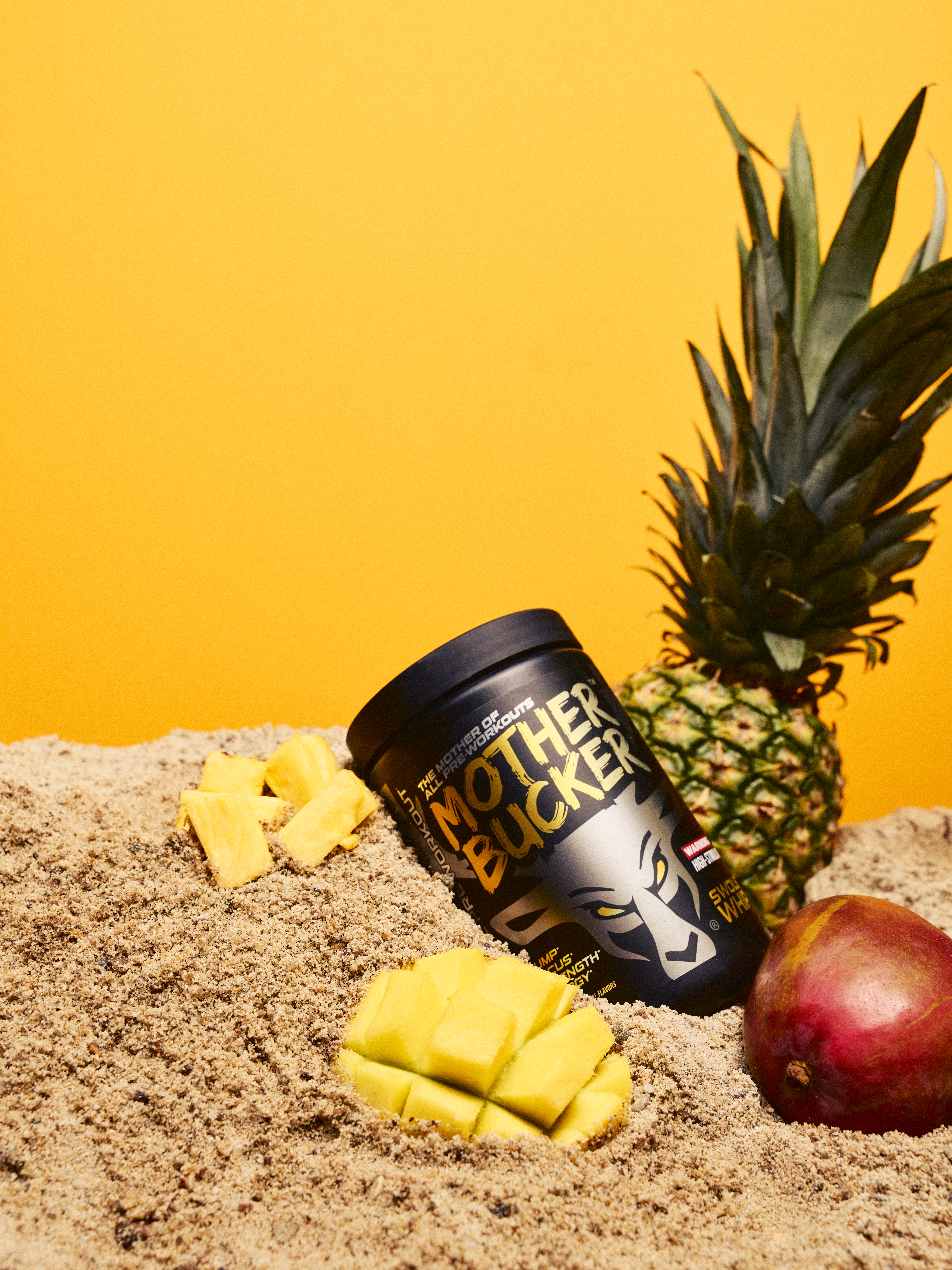 A black container of Mother Buckets protein powder is partially buried in light brown sand, with chunks of fresh pineapple placed around it. A ripe pineapple with spiky green leaves stands upright behind the container, and a red apple with a yellowish hue rests on the sand to the right. The background is a solid bright yellow, creating a vibrant, tropical-themed scene with high contrast and sharp focus, shot in a professional style with natural lighting highlighting the textures of the fruits and sand.