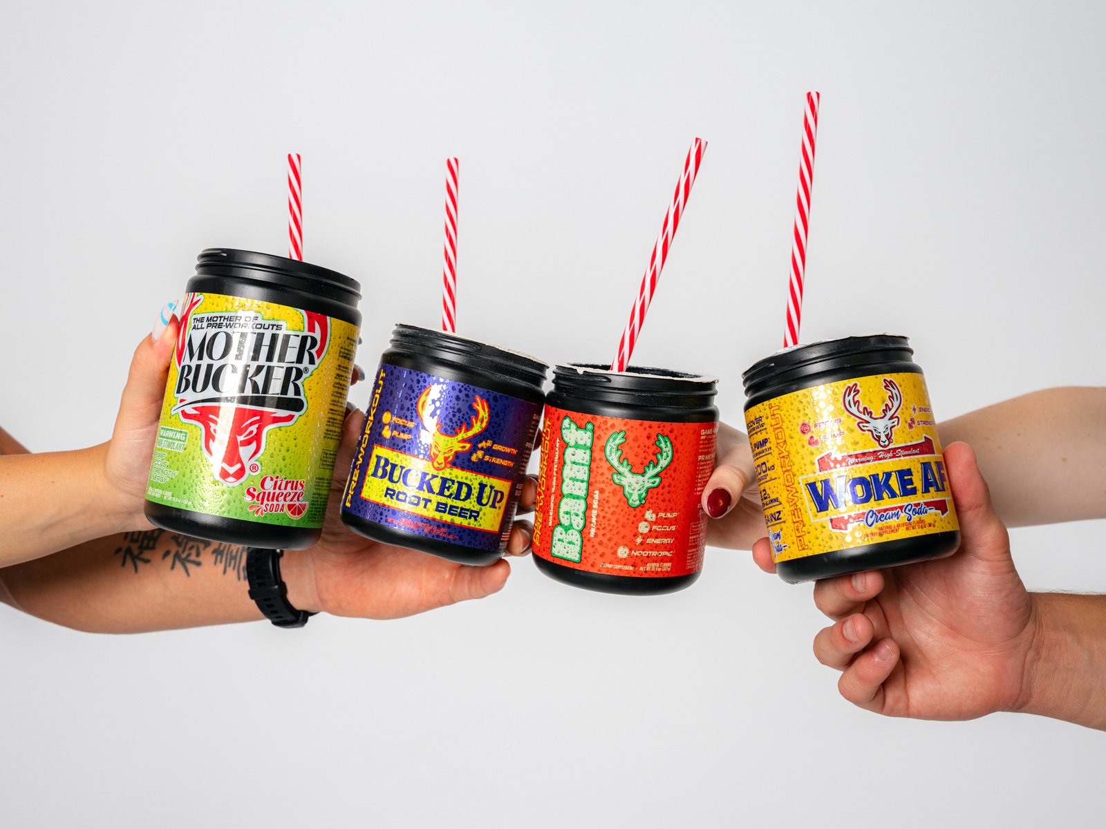 Four hands hold brightly colored Bucked Up supplement containers against a clean white background, each fitted with a red-and-white striped straw as if turned into fun, drinkable mocktails. From left to right: Mother Bucker (Citrus flavor) in a neon green label, Bucked Up (Root Beer flavor) with a metallic purple label, BAMF (oraflavor) in bold red, and Woke AF in yellow-gold. The arrangement and vivid design emphasize the playful yet powerful energy of the Bucked Up pre-workout line — a fusion of fitness, flavor, and lifestyle attitude.