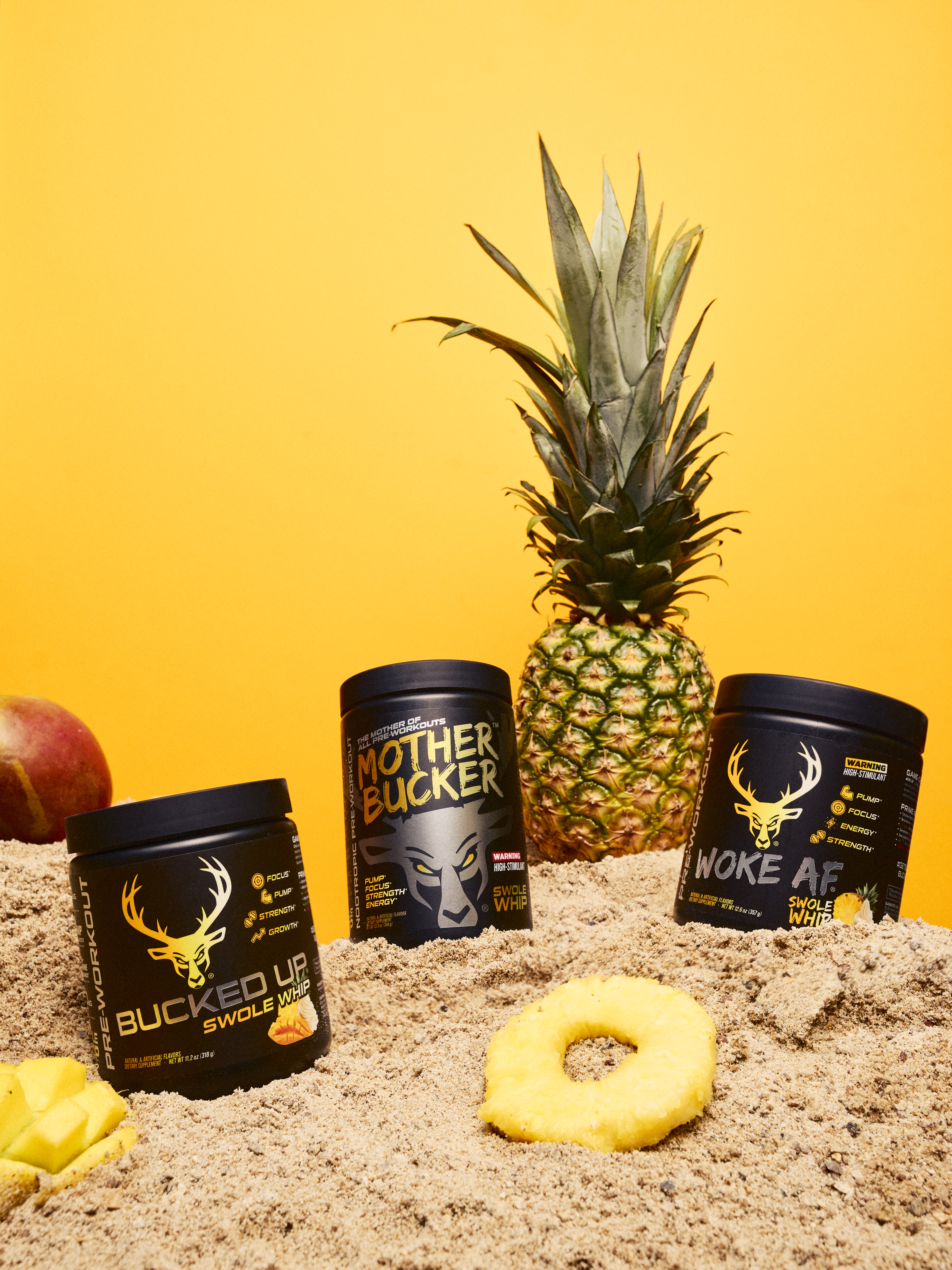 A vibrant display of three black containers of pre-workout supplements with bold yellow and white text and logos, arranged on sandy ground with a fresh pineapple standing upright behind them, a red apple partially visible on the left, and a yellow pineapple ring in the foreground, all set against a bright yellow background with soft lighting that highlights the textures of the sand, fruits, and containers.