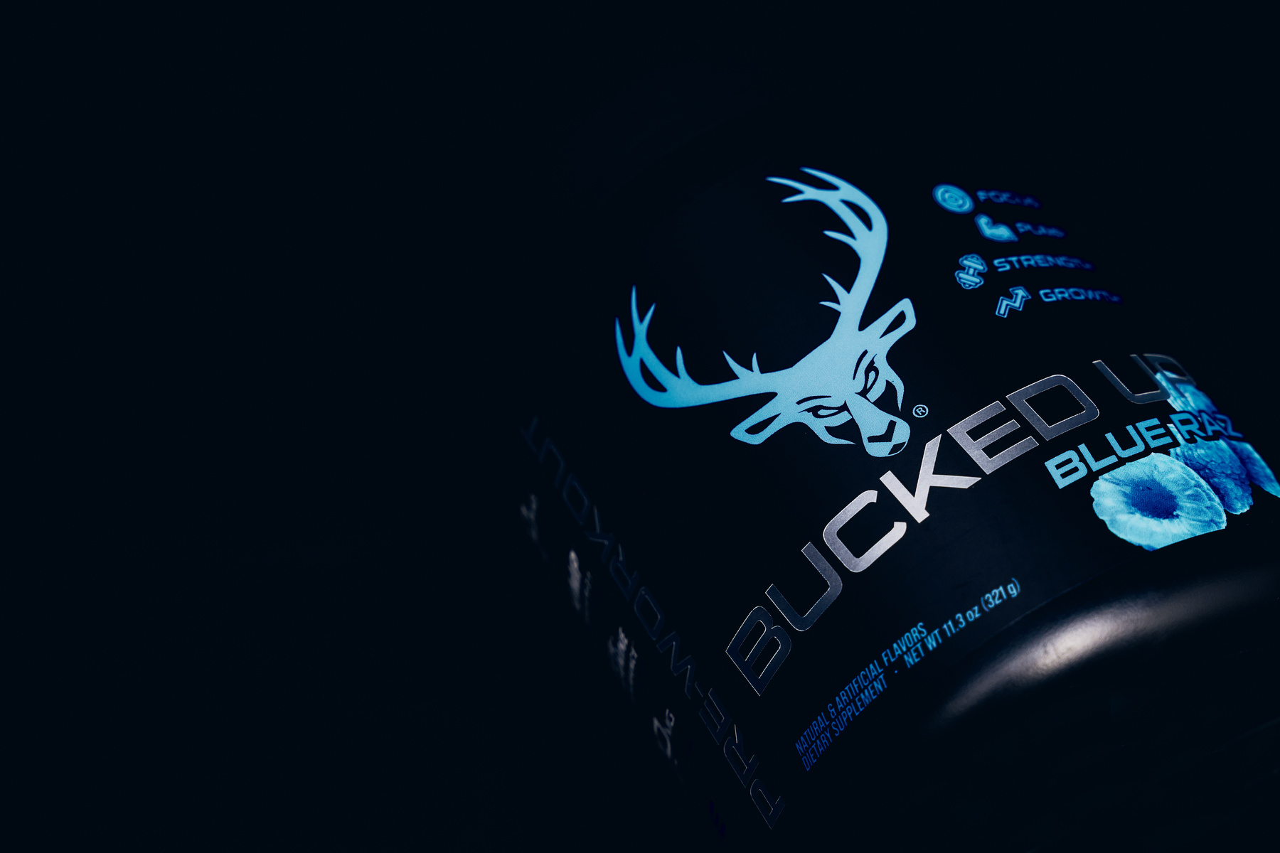 A black container of Bucked Up Blue Razz dietary supplement with a prominent white logo of a stylized deer head with antlers, illuminated by a soft blue light that highlights the product name and flavor. The label features icons representing focus, power, strength, and growth, arranged vertically on the right side, with the flavor image of a blue raspberry at the bottom. The overall mood is sleek and modern, with a dark background emphasizing the glowing elements and metallic text, suggesting a high-energy, performance-oriented product.
