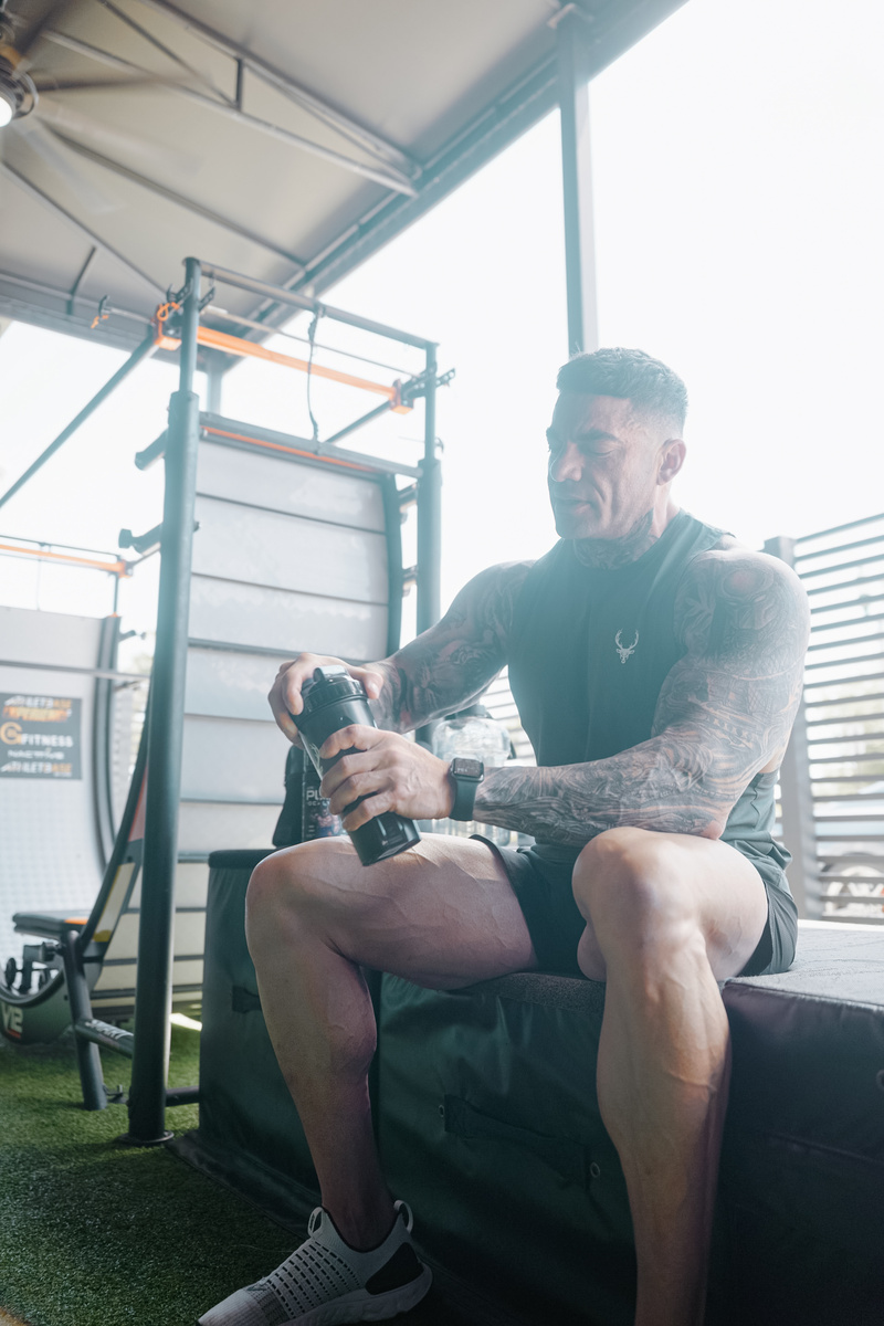“Muscular man in Bucked Up sleeveless workout shirt mixing pre-workout supplement in gym – fitness lifestyle photo promoting Bucked Up performance apparel.”