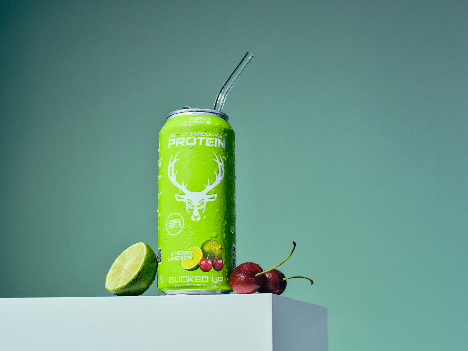 A bright green can of lightly carbonated protein drink with a white deer logo and text, placed on a white surface against a plain, muted green background. The can features a straw protruding from the top, with droplets of condensation visible on its surface. Next to the can, there is a halved lime with a vibrant green interior, and two red cherries with stems, one with a slight blemish. The composition is simple and clean, with soft, even lighting highlighting the freshness of the fruits and the sleek design of the can, creating a fresh, energetic mood suitable for a health or fitness brand.
