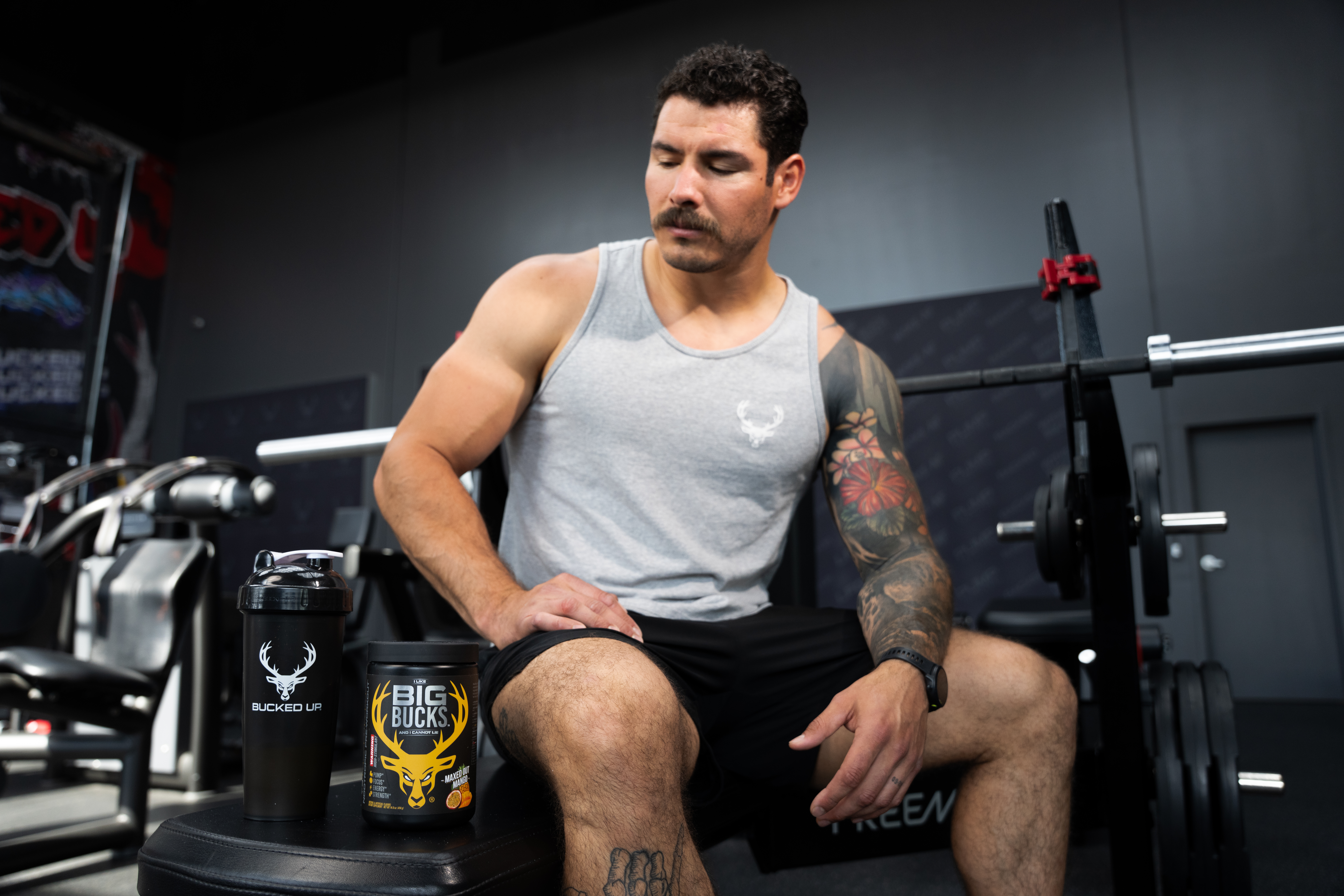 A muscular man with short curly dark hair, a mustache, and light skin, wearing a light gray sleeveless athletic shirt with a small white logo on the chest, is sitting on a black workout bench in a gym. He has a colorful floral tattoo sleeve on his right arm and a tattoo on his left hand. His expression is focused as he looks down, with his right hand resting on his thigh. In front of him on the bench are two black containers: a shaker bottle with a white deer logo and the words "BUCKED UP," and a tub labeled "BIG BUCKS" with a yellow deer head logo and flavor description. The gym background features dark walls, workout equipment, and a barbell with weights behind him, illuminated by bright, directional lighting that highlights his physique and the items on the bench.