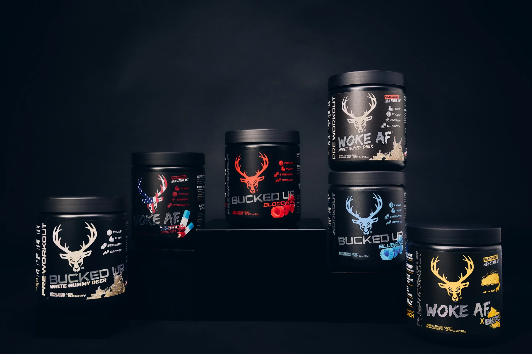 A collection of five black containers of dietary supplements with a consistent branding theme featuring a stylized deer head logo and bold text, arranged on a black platform against a dark background. The containers display different product names and flavors, with vibrant color accents such as white, red, blue, and yellow, and include icons indicating their benefits like focus, pump, energy, strength, and growth. The lighting highlights the containers' labels, creating a sleek, professional, and energetic aesthetic.
