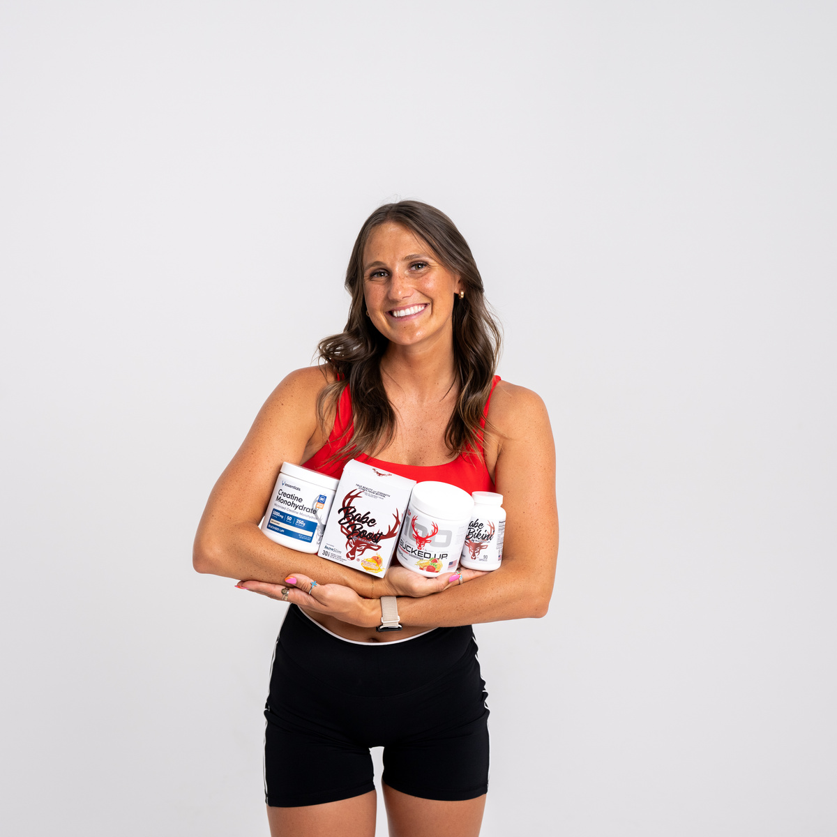 A smiling woman with long wavy brown hair, wearing a red sleeveless top and black pants, stands against a plain white background. She holds several health supplement bottles and boxes featuring a red deer logo and bold text, cradling them in her arms. The lighting is bright and even, highlighting her cheerful expression and the product details. The overall mood is energetic and positive, suitable for a health or fitness promotional image.