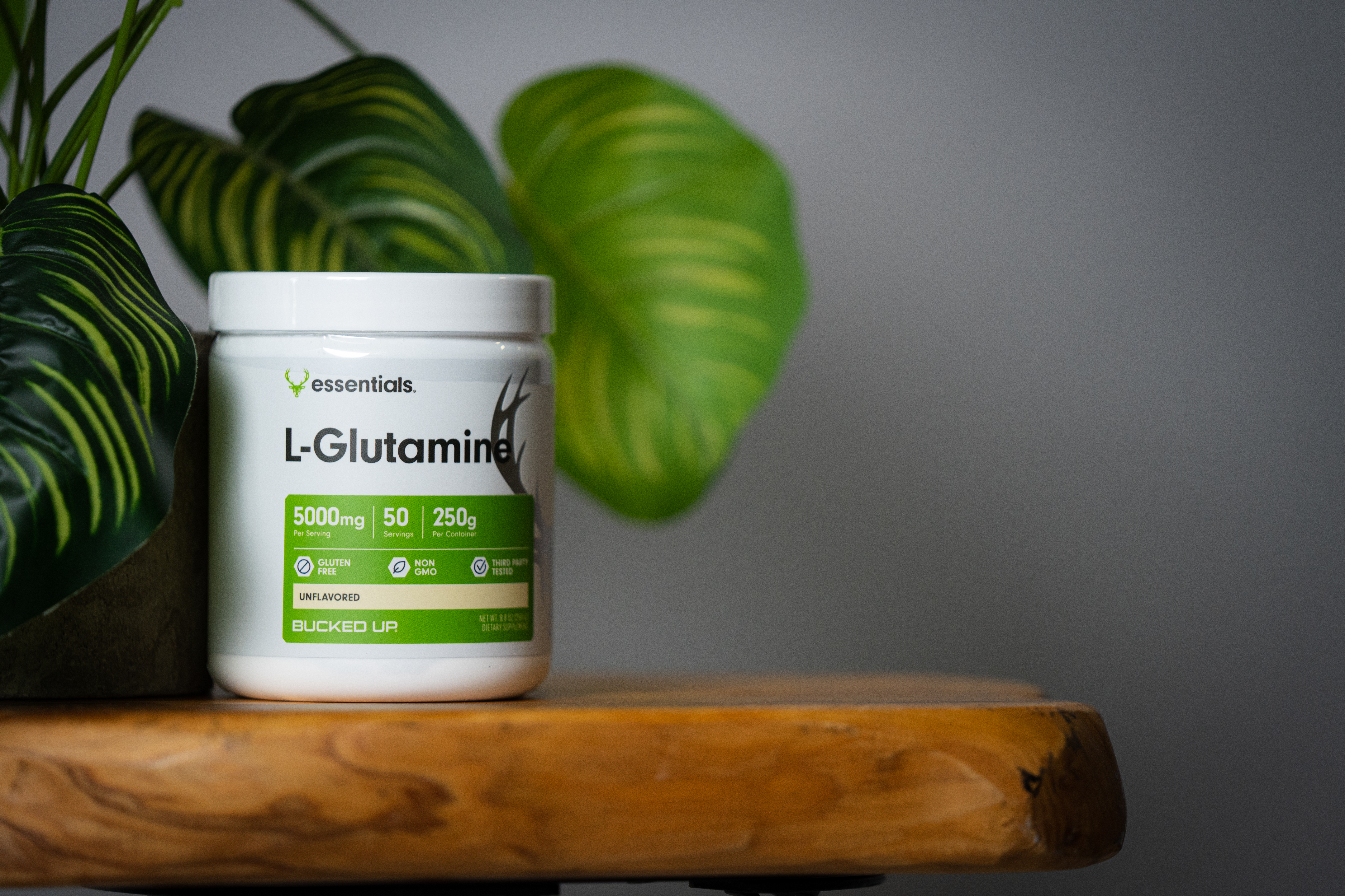 Bucked Up Essentials L-Glutamine supplement jar labeled '5000mg,' '50 servings,' and '250g,' gluten-free and non-GMO, set beside vibrant green leaves on a wooden surface.