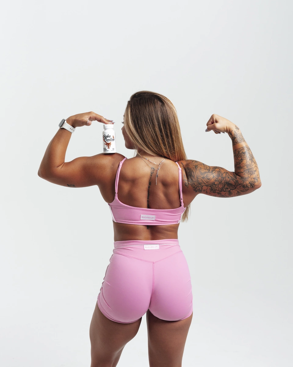 A muscular woman with long brown hair, tattoos on her right arm and back, wearing a pink sports bra and matching pink shorts, is standing against a plain white background. She is flexing her right arm to show her bicep and holding a white supplement bottle labeled 'Babe Creatine' on her left shoulder, with her face turned away from the camera. The lighting is bright and even, emphasizing her toned physique and the vibrant pink outfit.