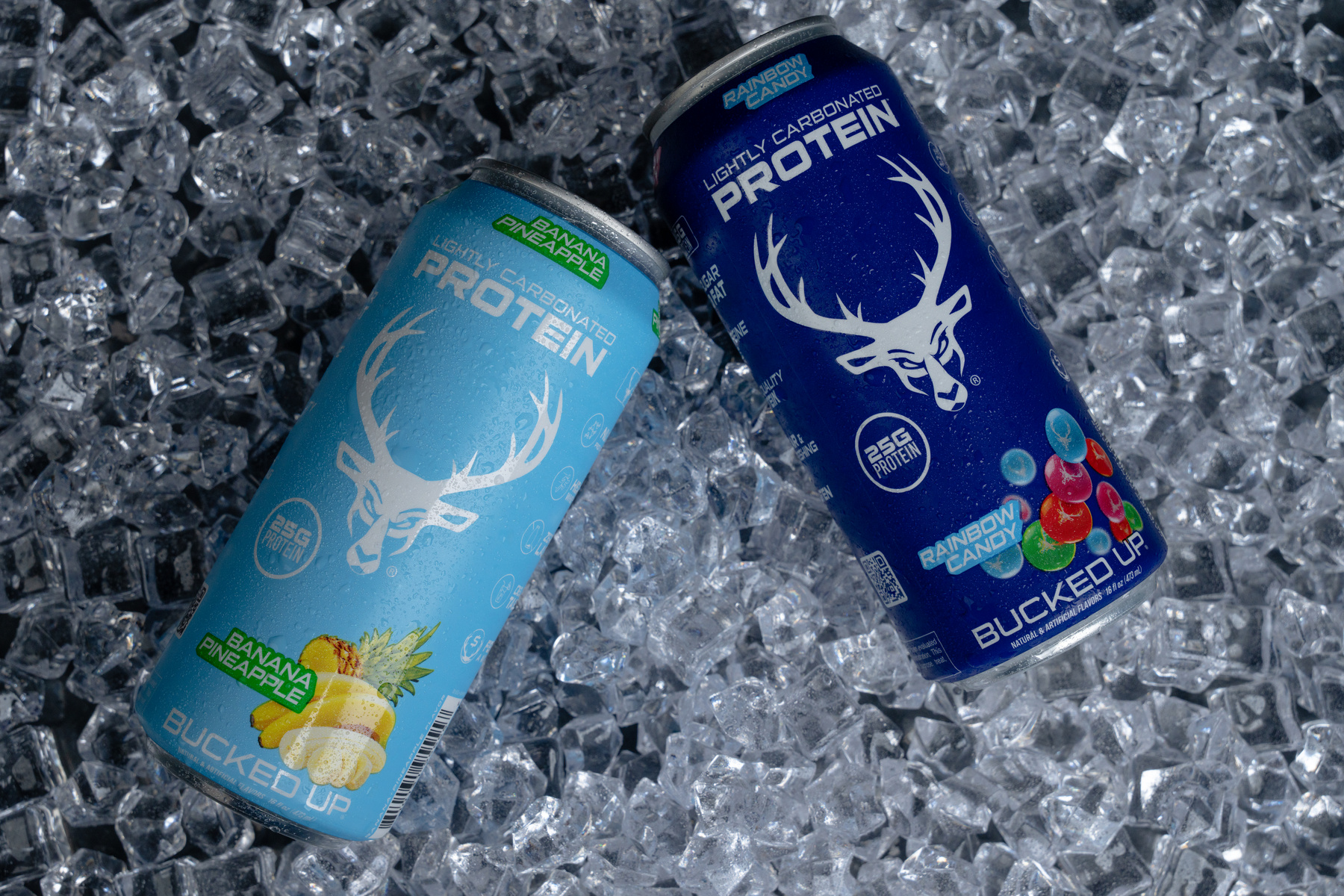 Two cans of Rainbow Candy lightly carbonated protein drink are placed on a bed of crushed ice. The can on the left is light blue with a pineapple and banana illustration, labeled "Banana Pineapple" flavor, featuring a white deer head logo and green accents. The can on the right is dark blue with a white deer head logo, labeled "Buck'ed Up" with rainbow-colored candy graphics, and indicates 25g of protein. Both cans are slightly tilted, with condensation droplets visible on their surfaces, and the background consists of clear, irregularly shaped ice pieces.