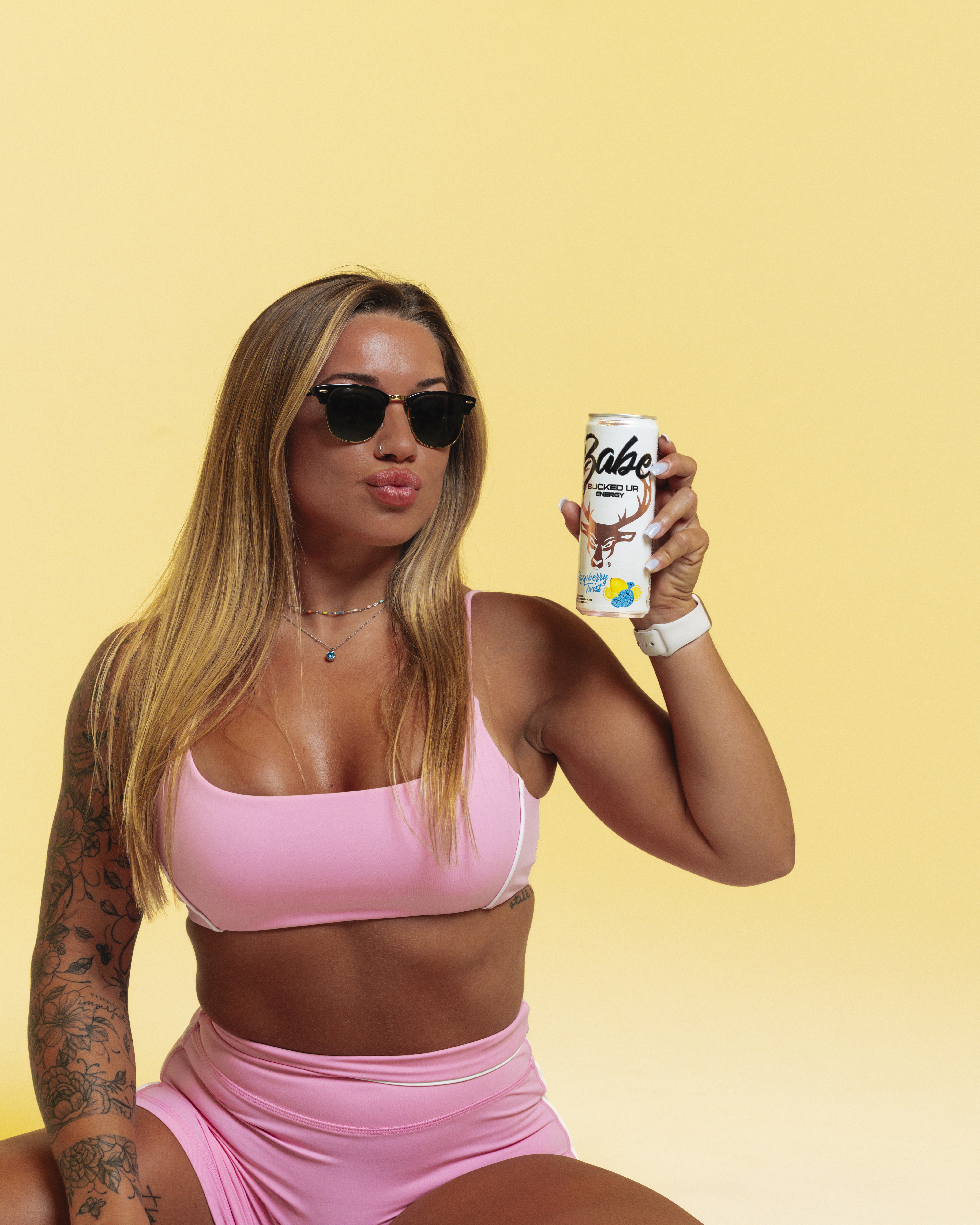 A young woman with long, wavy blonde hair wearing black sunglasses, a pink sports bra, and matching pink shorts, holding a can of energy drink labeled 'Babe Bucked Up' with a deer logo and colorful stickers, sitting against a plain yellow background, with a confident expression and puckered lips, styled in a casual, sporty look with minimal jewelry and visible tattoos on her left arm.