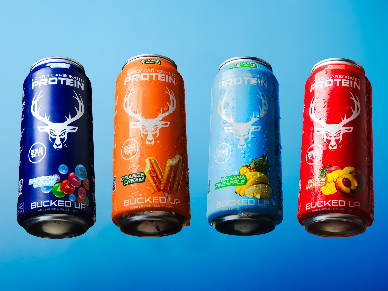 Four colorful cans of protein drinks are suspended in mid-air against a bright blue sky, arranged in a straight line with equal spacing. Each can features a sleek, modern design with a prominent white deer head logo, the flavor name, and vibrant fruit illustrations: rainbow candy, orange cream, banana pineapple, and peach mango. The cans are slightly wet with condensation, emphasizing freshness, and are illuminated by natural sunlight, creating subtle highlights and shadows that enhance their glossy finish. The overall mood is energetic and appealing, suitable for a health and fitness lifestyle advertisement.