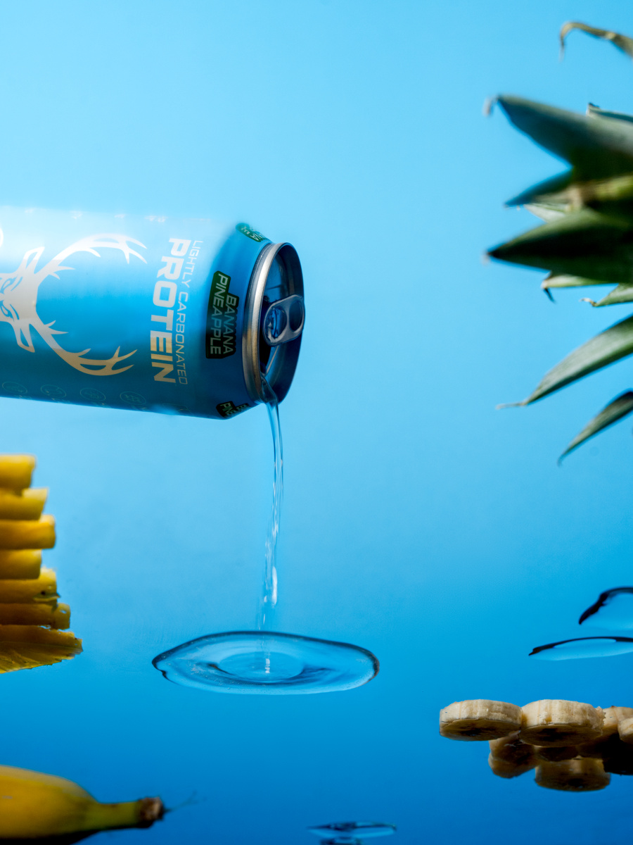 A blue aluminum can of lightly carbonated pineapple banana flavored beverage is tilted in mid-air, pouring a stream of clear liquid into a reflective surface below, creating a ripple. The can features a white stylized deer logo and text, with a green tab open. The background is a solid bright blue, with parts of a pineapple with spiky green leaves on the right, and slices of banana and pineapple on the left and bottom, arranged around the can. The scene is brightly lit with soft shadows, emphasizing the vibrant colors and glossy textures, styled as a modern product shot.