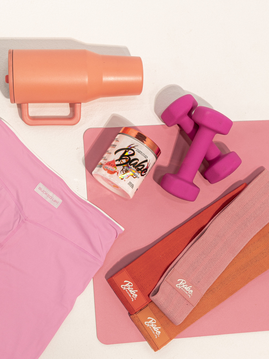 A flat lay arrangement of fitness and wellness items on a pink and white background, including a pink water bottle with a handle, a pink dumbbell, a container of Bude protein powder with a colorful label, a pink yoga mat partially rolled up, and two folded yoga towels in pink and orange with the Bude logo, all illuminated with soft, even lighting creating subtle shadows and highlighting the vibrant pastel color palette, styled in a modern, energetic lifestyle aesthetic.