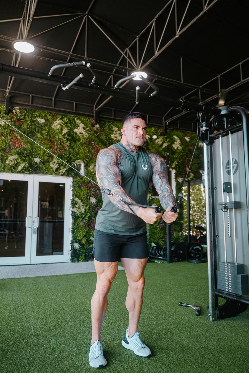 “Tattooed man performing cable chest fly exercise wearing Bucked Up sleeveless training shirt and black shorts at indoor gym – fitness apparel and workout lifestyle photo.”
