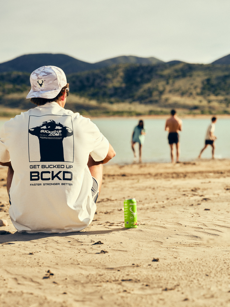 A young man with light skin, wearing a white t-shirt with black graphics and text, and a camouflage baseball cap with a deer logo, is sitting on sandy beach facing away from the camera. He is looking towards two people walking into the water, one in a light blue shirt and the other shirtless in black shorts. The background features a lake with calm water, green hills, and mountains under a clear sky. A bright green canned beverage with a deer logo is placed on the sand near him. The scene is lit by warm, natural sunlight, creating soft shadows and highlights, with a relaxed, outdoor summer mood.