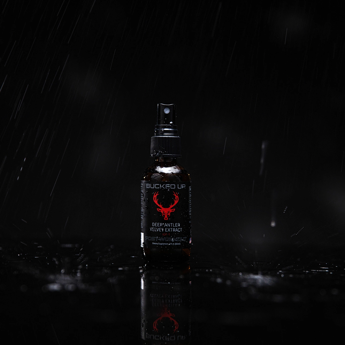 A small dark amber spray bottle with a black spray nozzle stands upright on a wet, reflective black surface, with water droplets scattered around its base. The bottle features a black label with a red deer antler logo and white text that reads "BUCKED UP" and "DEER-ANTLER VELVET EXTRACT." The background is completely black, emphasizing the bottle and its reflection, with subtle highlights on the water droplets and the bottle's surface. The lighting is focused from above, creating a slight sheen on the bottle and casting soft shadows, conveying a sleek, professional, and slightly mysterious mood.