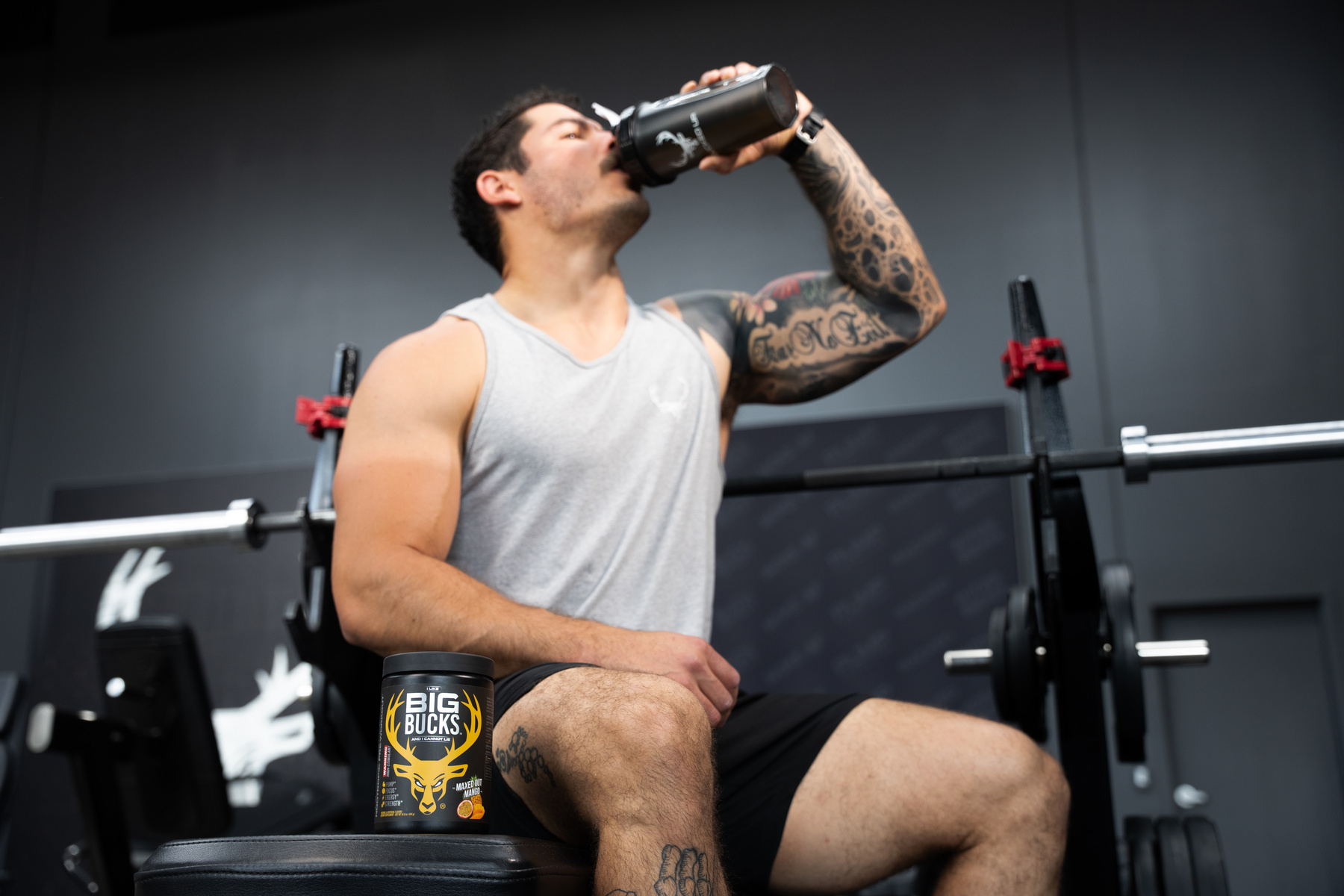 A muscular man with tattoos on his arms, wearing a light gray sleeveless shirt and black shorts, is sitting on a workout bench in a gym, drinking from a black shaker bottle. The gym has a dark gray wall with a white logo of a deer head in the background, and a barbell with weights is positioned behind him. A container labeled 'Big Bucks' with a deer antler logo is placed on the bench next to him. The lighting is bright and focused, highlighting his physique and tattoos, creating a professional fitness environment.