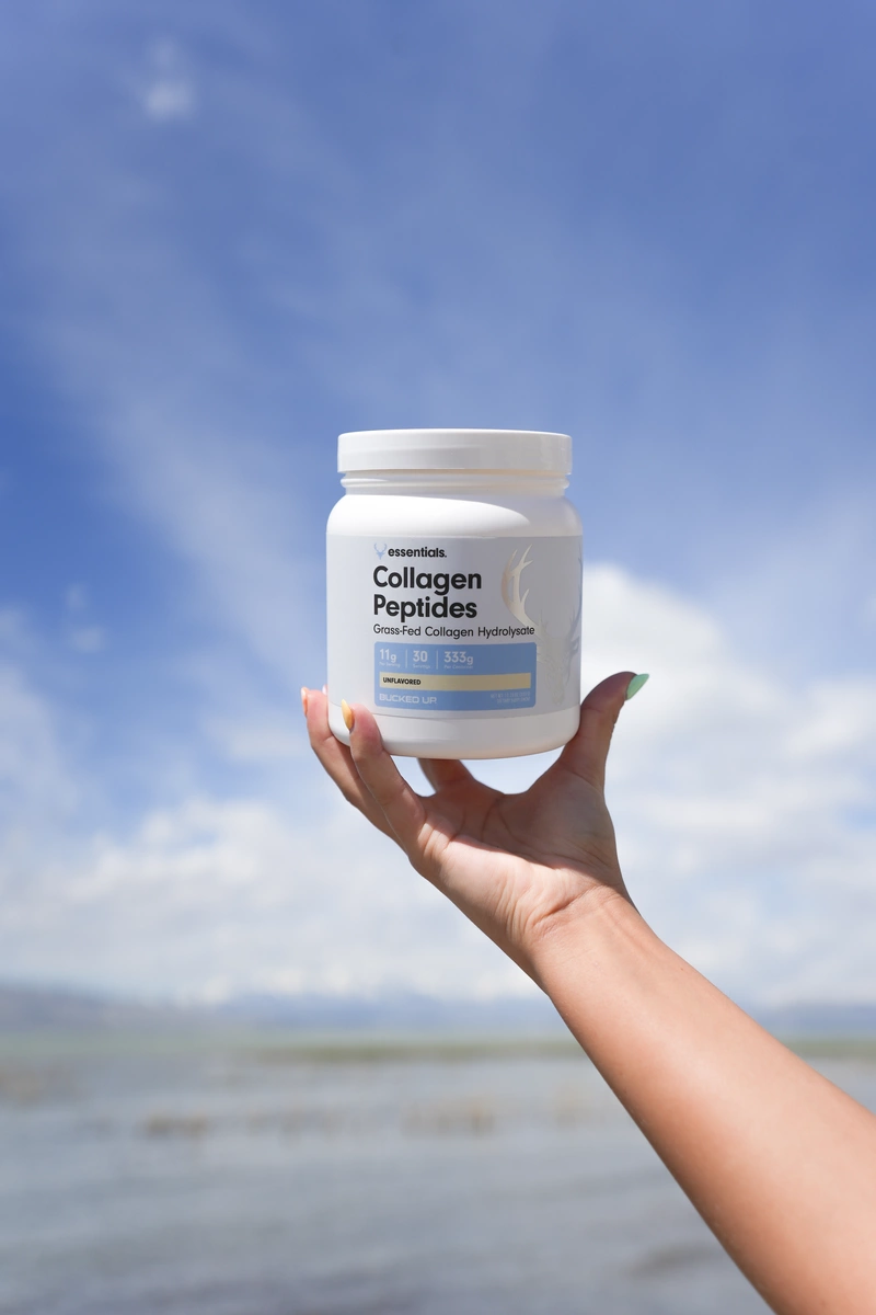 A hand with a light tan skin tone holds a white container of 'Collagen Peptides' against a bright blue sky with fluffy white clouds. The container features a clean design with a label that includes the text 'Grass-Fed Collagen Hydrolysate' and nutritional information in black and yellow. The hand is positioned slightly above eye level, creating a dynamic angle that emphasizes the product. The background is a serene outdoor setting with hints of water and distant mountains, suggesting a natural and healthy lifestyle. The lighting is bright and sunny, casting soft shadows and highlighting the container's smooth surface, contributing to an uplifting and fresh mood.