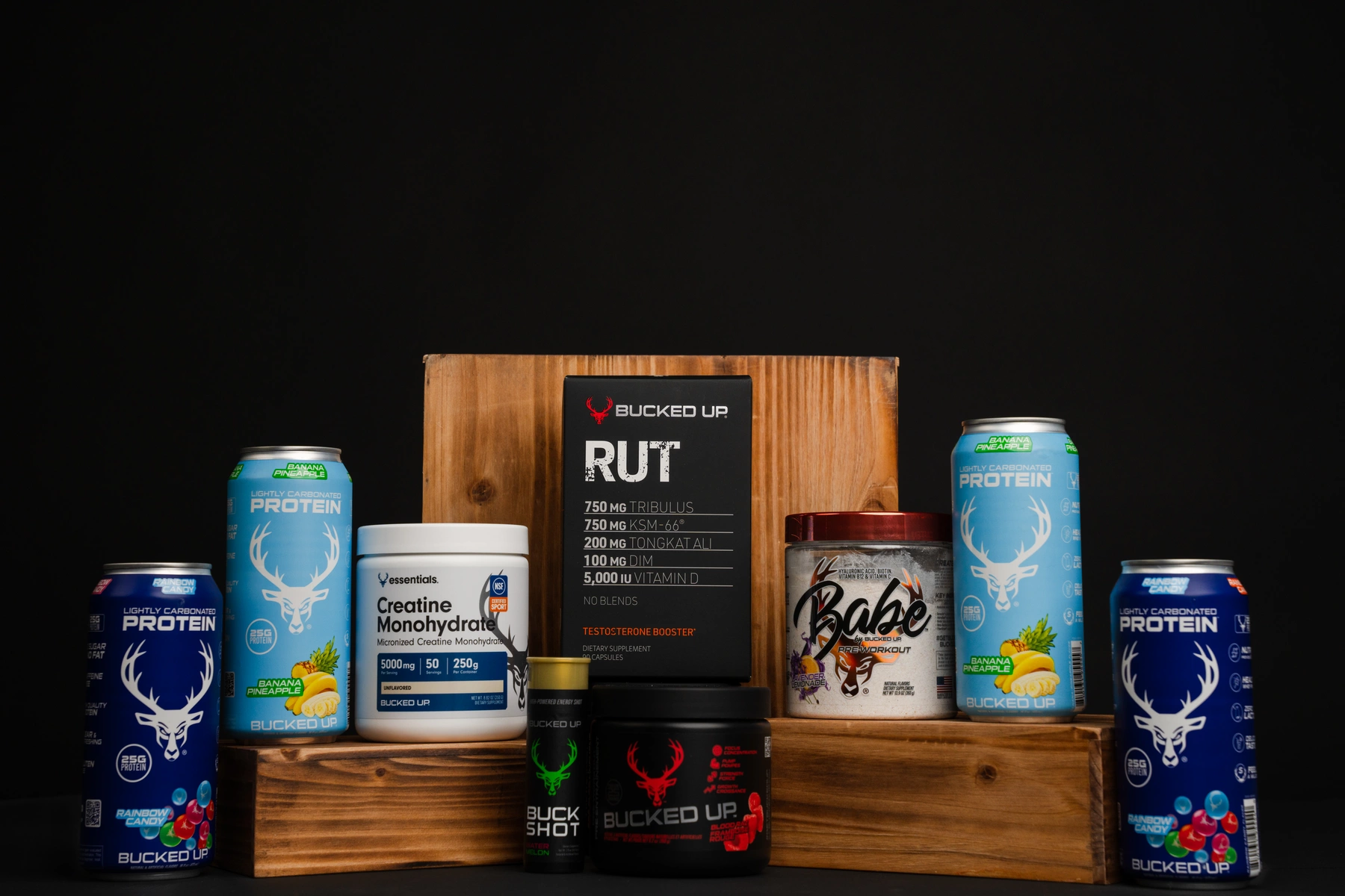 A collection of fitness supplements and protein powders arranged on a wooden platform against a black background, including three blue cans of lightly carbonated protein drinks with a white deer logo, a white container of creatine monohydrate with a blue label, a black and red testosterone booster box labeled 'RUT' with detailed ingredient information, a small black and green bottle labeled 'Buck Shot,' a red-lidded jar of pre-workout labeled 'Babe,' and a blue and white container of protein powder with a similar deer logo, all positioned to showcase their labels and branding, with soft, focused lighting highlighting the products' colors and textures.
