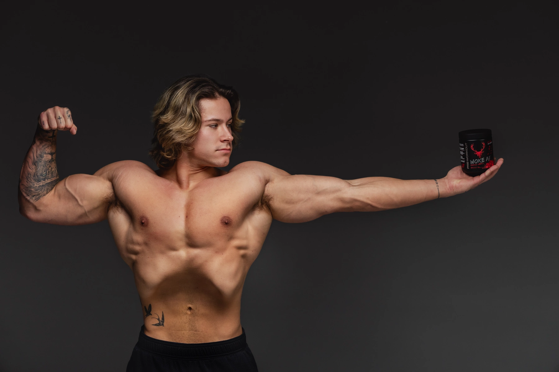 A muscular young man with shoulder-length wavy blond hair and light skin is shirtless, flexing his right bicep while holding a black container with red and white text and a red deer logo in his left hand, extended outward. He has tattoos on his right arm and left side of his torso, and a small tattoo on his right wrist. The background is plain dark gray, with soft, even lighting highlighting his physique and facial features, creating a professional, fitness-themed portrait.